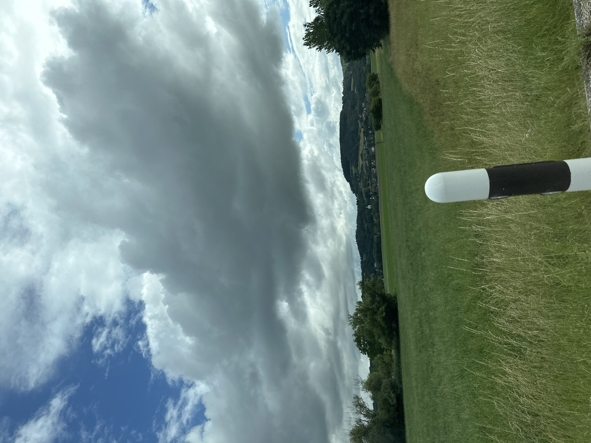 The image presents a wide, expansive landscape under a dynamic sky. In the foreground, prominently in the bottom right, a white cylindrical object with a rounded top is visible, featuring a distinct black band around its middle section, possibly a post or marker. The immediate ground consists of varied green grass, with some areas appearing longer and drier.
Beyond the foreground, a large, lush green field or meadow stretches towards a distant line of trees and hills. These hills are densely covered in dark green foliage, with hints of scattered buildings or houses nestled within, suggesting a rural or semi-rural settlement.
The sky occupies a significant portion of the upper frame and is marked by dramatic cloud formations. Large, heavy, dark grey clouds dominate much of the sky, giving an impression of overcast or possibly stormy weather. However, patches of bright blue sky are visible, particularly in the lower left and upper right corners, interspersed with lighter, wispy white clouds, indicating a changing weather pattern.
The scene captures a daytime moment with no people or specific activities taking place, focusing instead on the natural environment. The rolling, tree-covered hills and green fields under a compelling sky are consistent with the scenic landscapes found in areas like Zurzach, Switzerland. No visible text is present in the image.