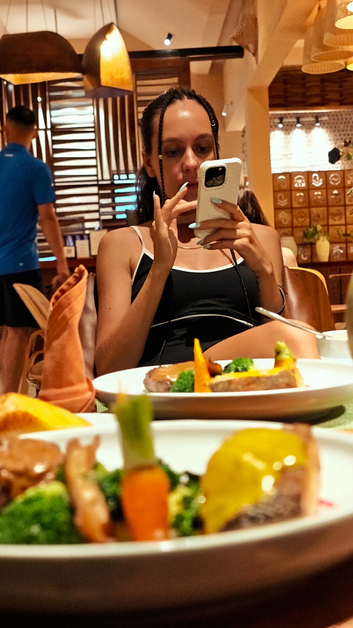 The image shows a young woman with braided hair seated at a table in a restaurant, looking at her phone. She is wearing a black tank top with white trim. In front of her are plates of food, including what appears to be steak, broccoli, and carrots. An orange napkin is folded decoratively on the table. The restaurant appears to have a warm, ambient lighting. In the background, another person in a blue shirt is visible, and decorative elements of the restaurant's interior are present.