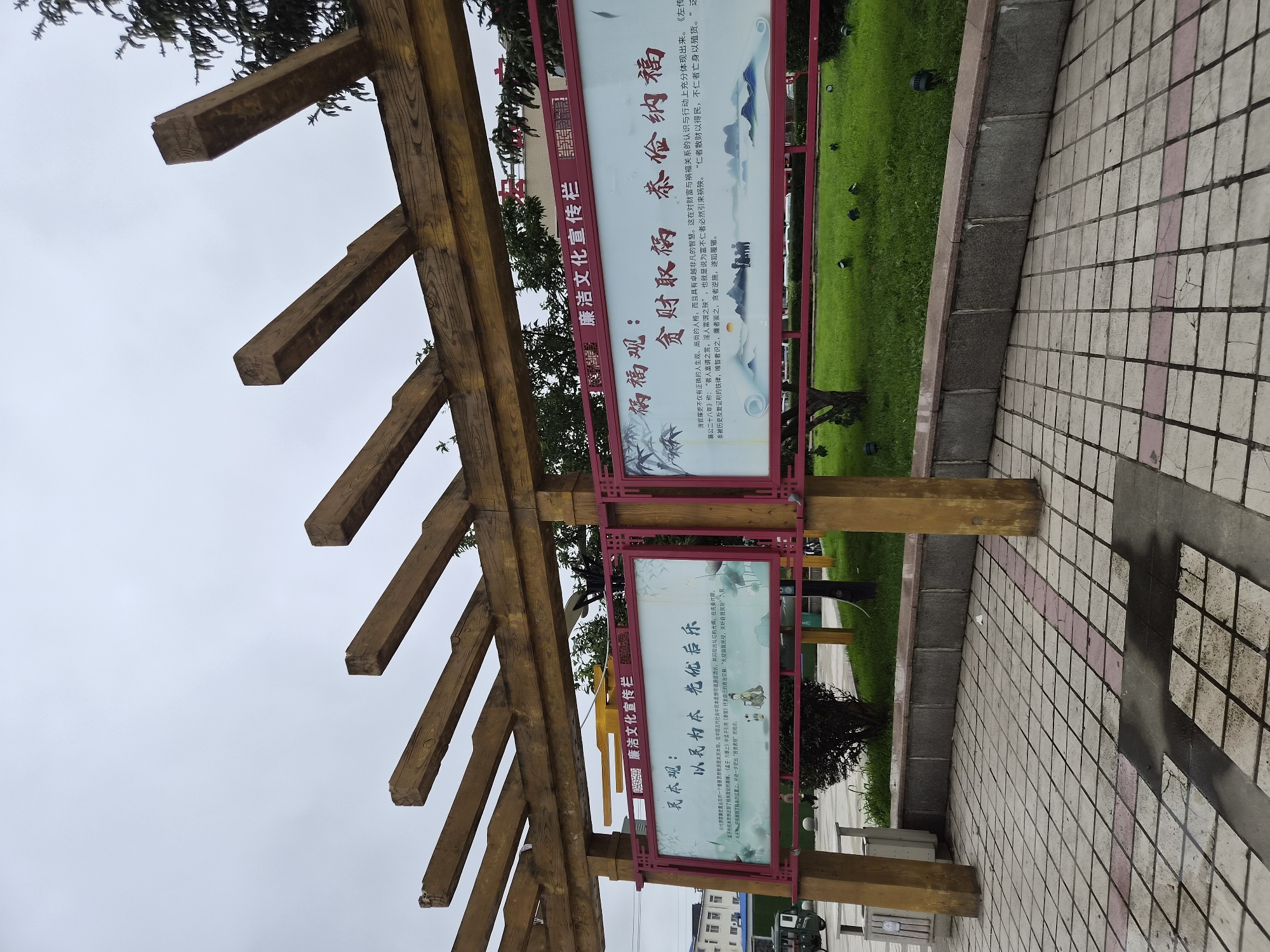 In the city of Hunchun, China, an outdoor scene features a wooden pergola structure with two large, rectangular display boards. The left board, with a light background and red border, contains Chinese text and an illustration depicting lotus flowers and a small dog. The right board, also with a light background and red border, has Chinese text and a landscape illustration of mountains, a body of water, and silhouetted figures. Both boards are mounted on yellow-painted pillars. In the background, a well-maintained green lawn is visible, dotted with several dark outdoor lights. Beyond the lawn, trees and a building with a red sign can be seen. To the left of the pergola, a small, three-wheeled vehicle and a trash receptacle are present. The ground in the foreground is paved with square tiles, some of which are wet, indicating recent rain or a puddle. The sky is overcast and gray.