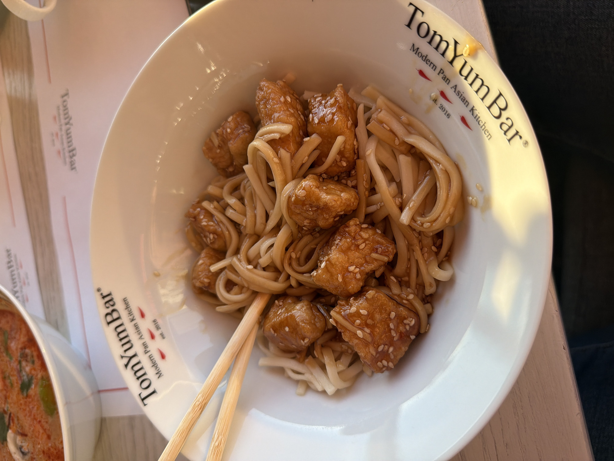 The image shows a white bowl filled with noodles and cubed, sesame-covered pieces, likely chicken or tofu, served at Tom Yum Bar, a modern Pan-Asian kitchen established in 2016, as indicated by the text on the bowl and adjacent menu. A pair of chopsticks rests on the edge of the bowl. A smaller white bowl containing a reddish-orange broth-based soup is partially visible in the lower left corner. The setting is a restaurant, with a wooden table surface partially visible and a person in dark clothing to the right of the bowl. While the text mentions Tom Yum Bar, and the picture shows a Tom Yum soup in the background, the location of the restaurant (Krasnoyarsk, Russia) is not discernible from the image itself.