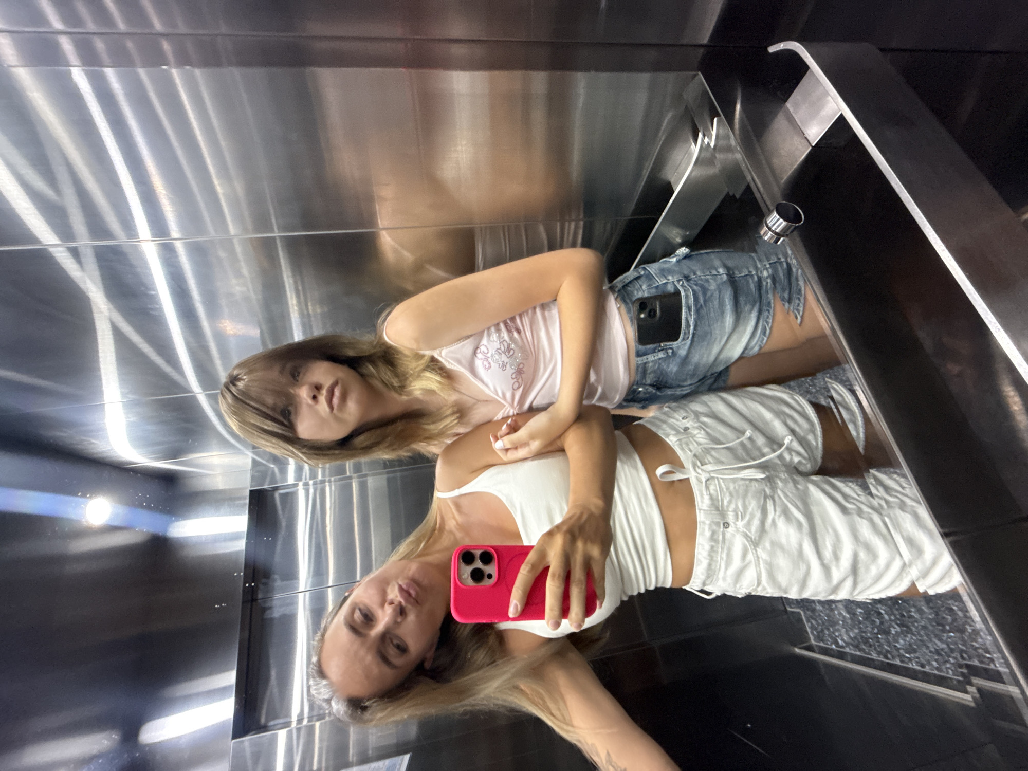 This is a mirrored selfie taken in what appears to be an elevator. Two young women are the subjects, with their reflections also visible. The woman in the foreground is wearing a white tank top and white shorts. She is holding a bright pink smartphone to take the picture. Her hair is long and blonde, and she has a relaxed expression. The woman behind her is wearing a pink camisole with floral embellishments and denim shorts. She has blonde hair as well and is looking towards the camera. The elevator walls are made of brushed metal, creating reflections and highlights that contribute to the overall aesthetic of the image. The lighting suggests it is daytime, but there are no specific cues about the weather or precise time of day. The location is identified as Khok Kham Subdistrict, Thailand, though this context isn't directly evident from the visual elements within the image itself. The overall mood is casual and playful, typical of a selfie taken in an everyday setting.