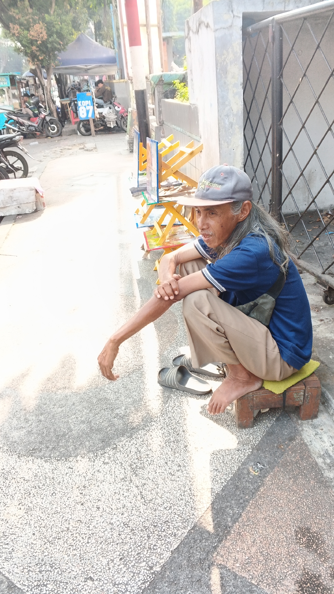 An elderly man with long, gray hair and a mustache sits on a wooden stool topped with a yellow cloth, facing left on a textured concrete sidewalk in Malang, Indonesia. He is wearing a blue polo shirt with white stripes on the collar and sleeves, tan pants, a gray baseball cap, and gray slide sandals. A dark green bag is slung across his chest. His left arm is resting on his lap, with his hand open and fingers splayed. The background features a street scene with several motorcycles parked, a blue tent with signage that includes "KOPI 87," and a metal fence. Sunlight casts shadows across the sidewalk.
