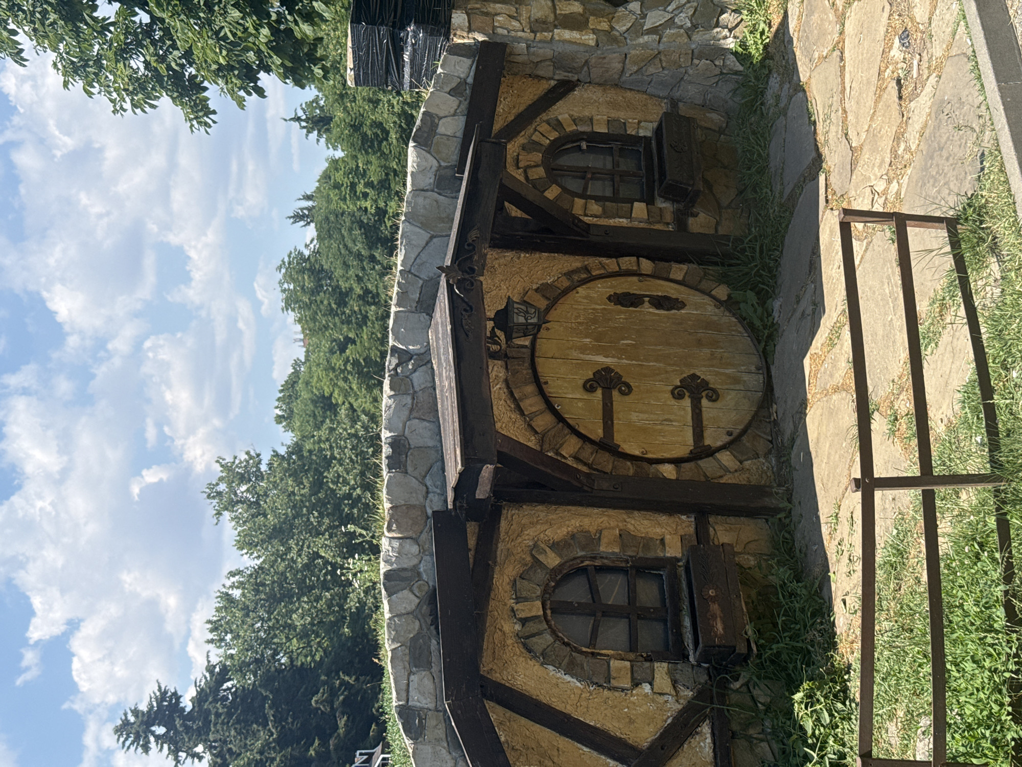 The image shows a whimsical, hobbit-style dwelling with a round wooden door and small, arched windows. The building is constructed with rough-hewn stone and plastered walls, accented with dark wooden beams and decorative metalwork. The style suggests a fantasy or fairytale theme.
The scene is set outdoors on a sunny day, with a bright blue sky filled with fluffy white clouds visible in the background, peeking through lush green trees. The ground in front of the dwelling is paved with irregular stone slabs, and thick green grass grows around the building and between the paving stones. A dark metal railing runs along the right side of the image, suggesting a pathway or an edge.
There are no people visible in the image, and no specific activity is taking place. The focus is entirely on the charming, storybook architecture of the dwelling and its natural surroundings. The overall impression is one of a peaceful, idyllic, and perhaps secluded location, likely a garden or a themed park. The light suggests it is daytime, possibly late morning or early afternoon, given the shadows cast by the building and the brightness of the sky.
The location context of Burgas, Bulgaria, implies this dwelling might be part of a resort, a private residence designed with a unique aesthetic, or a public attraction. No visible text is present in the image.