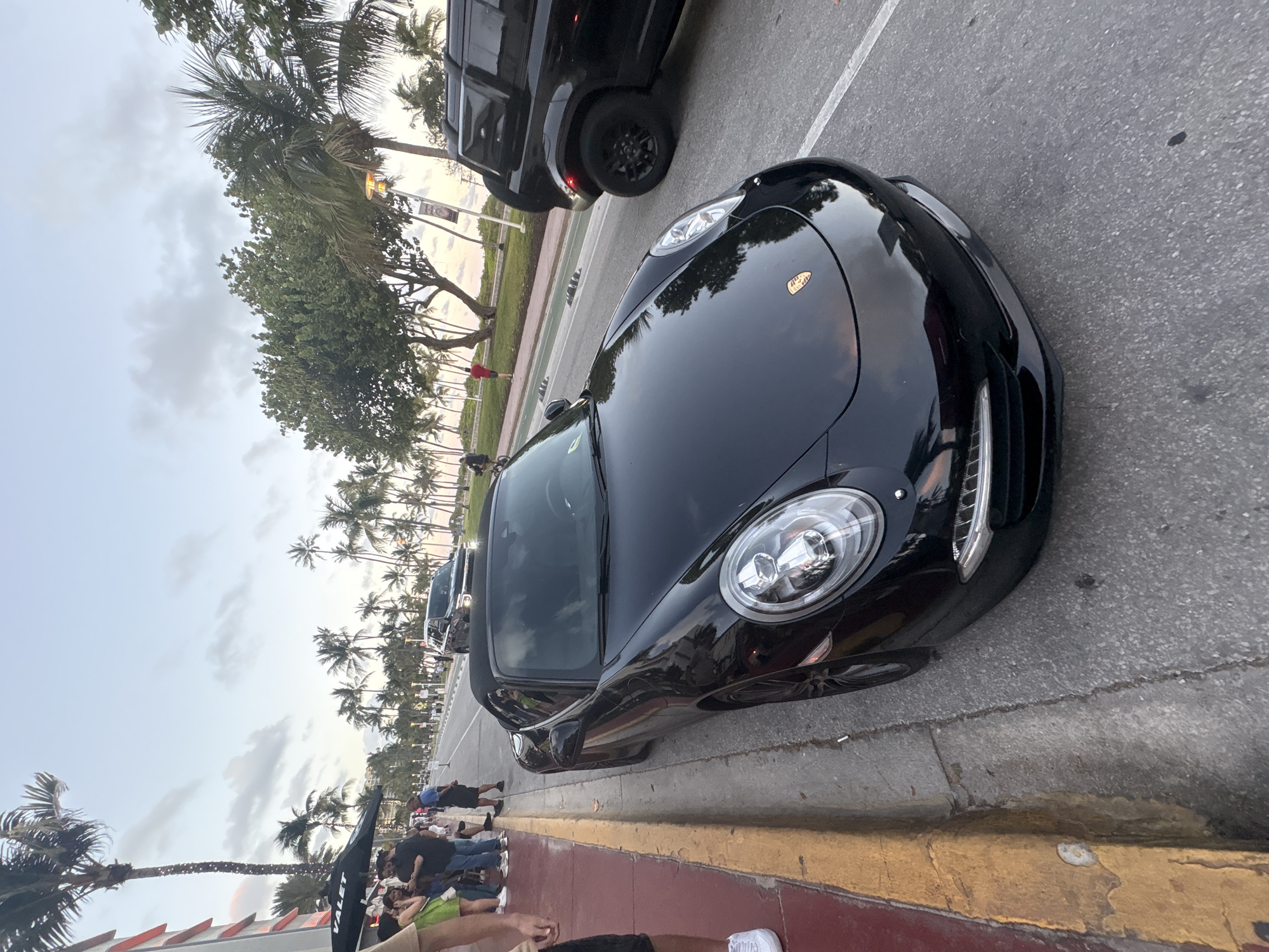 The image captures a street scene in Miami Beach, United States of America, likely during late afternoon or early evening due to the overcast sky. A sleek black Porsche convertible is prominently featured in the foreground, parked or stopped at an angle on the side of an asphalt road. Its front end, including the distinctive headlights and a Porsche emblem on the hood, is visible.
Behind the Porsche, another dark-colored SUV is also present on the road. A bright yellow curb separates the asphalt road from a reddish sidewalk. Along the sidewalk, numerous people are gathered in groups, some standing and others seemingly walking.
The background is dominated by lush tropical foliage, including numerous tall palm trees and other green trees, suggesting a scenic boulevard or a park adjacent to the street. A black umbrella with "VALET" written in white text is partially visible among the crowd on the sidewalk, hinting at a nearby service or establishment. The overall atmosphere appears casual and lively.