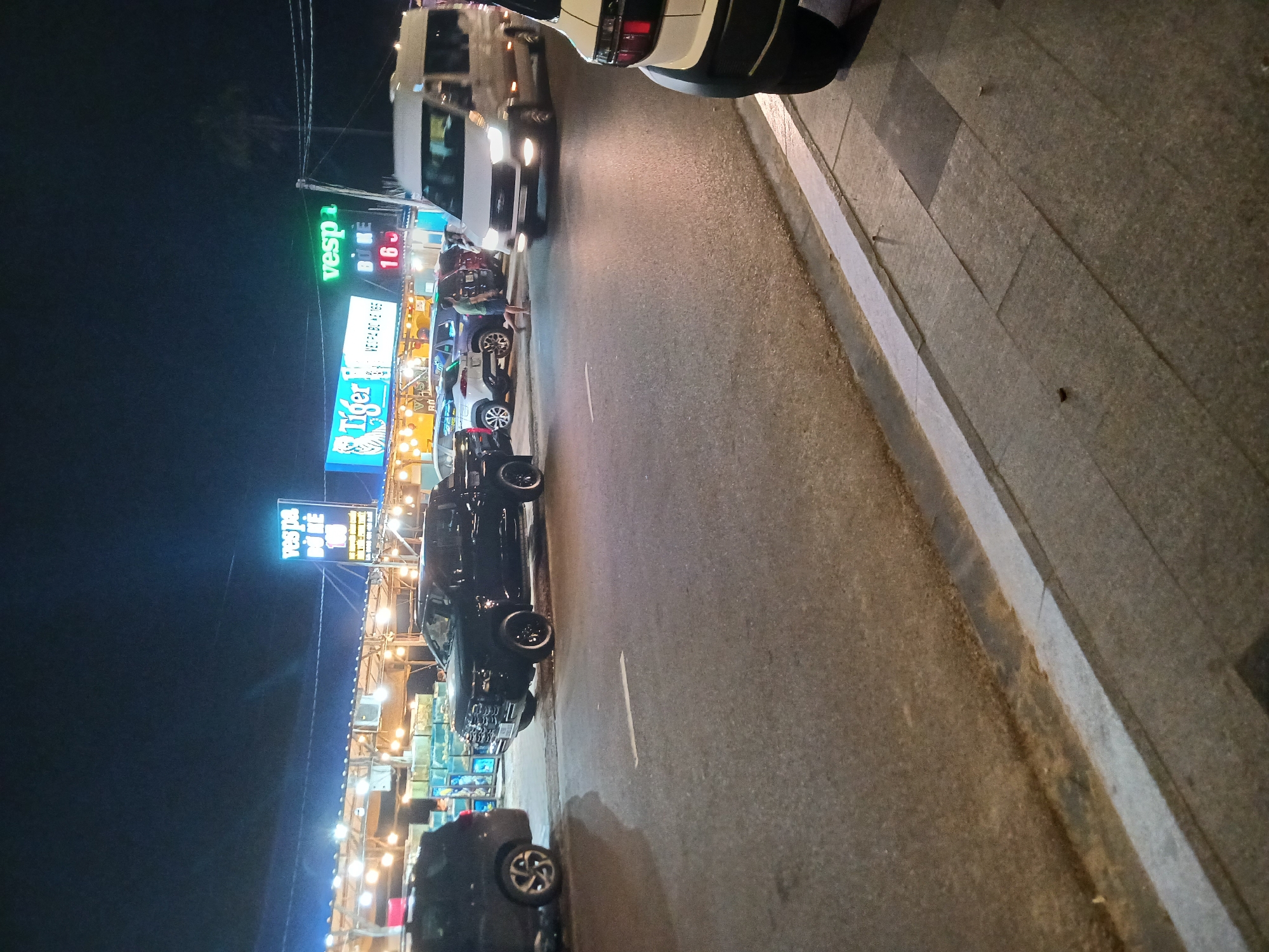 At night, a street in Phu Thuy Ward, Vietnam, features a row of establishments with bright neon signs. The signs display "Vespa" in blue and green, "Tiger" with a tiger graphic in blue and white, and "16" in red. Several vehicles are parked and in motion on the asphalt road, including a black pickup truck, a white van, and other cars. A person in a green shirt and blue shorts is walking on the road. A sidewalk with paving stones runs alongside the road, illuminated by overhead lights from the establishments. The sky is dark, indicating nighttime.