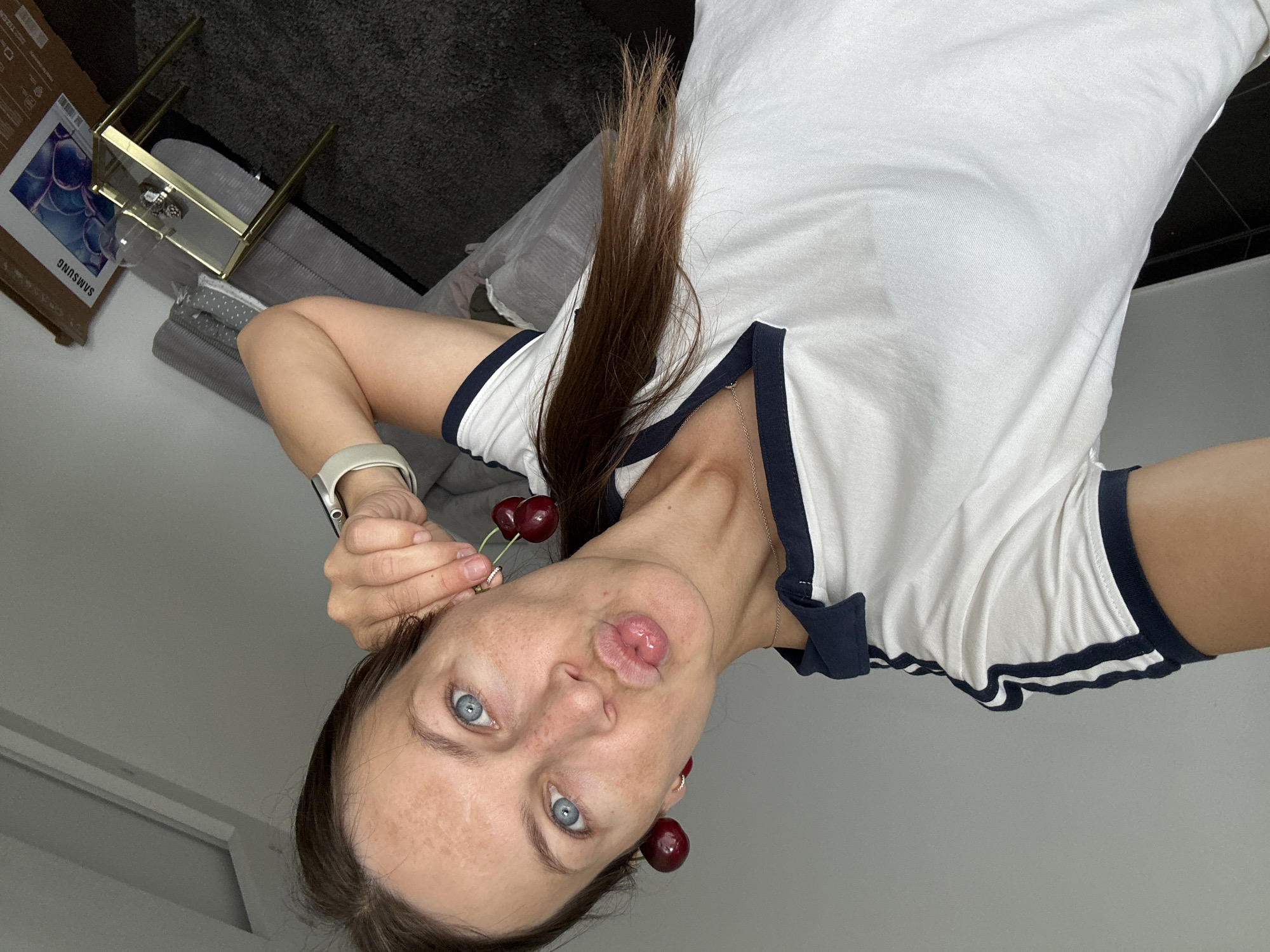 A young woman is taking a selfie indoors, with a neutral gray wall as the background. She is wearing a white polo shirt with navy blue trim. Her right ear has a cherry clipped to it, and she is holding two more cherries in her right hand, which is near her face. Her mouth is pursed as if she is blowing a kiss. She has blue eyes and brown hair tied back in a ponytail. A gray sofa and a dark gray carpet are visible in the background, along with a small gold side table with a glass top. A large cardboard box with the Samsung logo and a picture of a television is leaning against the wall to the right. The scene appears to be a domestic setting.