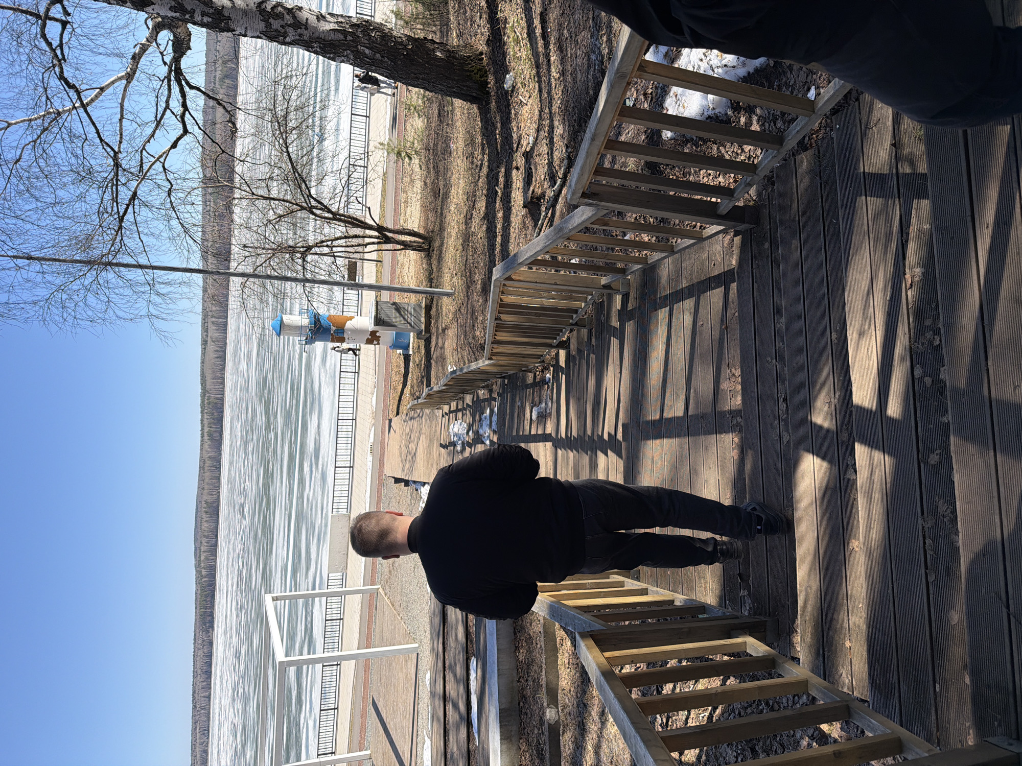 A man in black clothing stands on a wooden boardwalk with stairs and railings near the Kama River in the city of Chaikovsky, Russia. The river is partially covered with ice, and the sky is clear blue. A birch tree is visible on the left. The boardwalk leads down to a paved area with a white metal railing along the river.