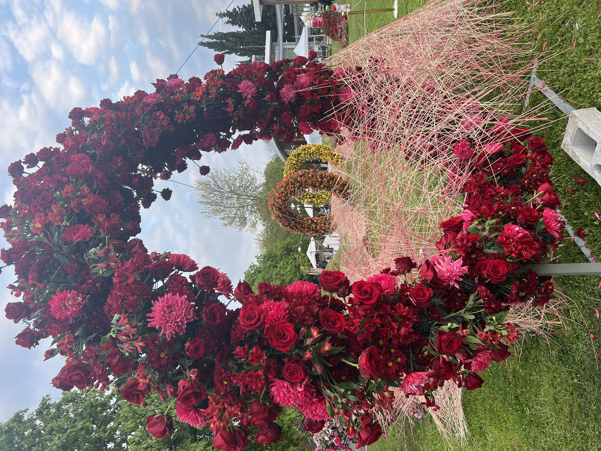 The image captures a close-up, slightly angled view of a circular floral arrangement, primarily composed of deep red roses and pink chrysanthemums. A light pink, interwoven structure of thin rods lies beside the flowers. A grassy area with scattered blooms surrounds the arrangement. In the blurred background, hints of white tents or canopies suggest an event or outdoor exhibition. Other circular floral displays are visible in the distance. This event likely took place in Burgas, Bulgaria.