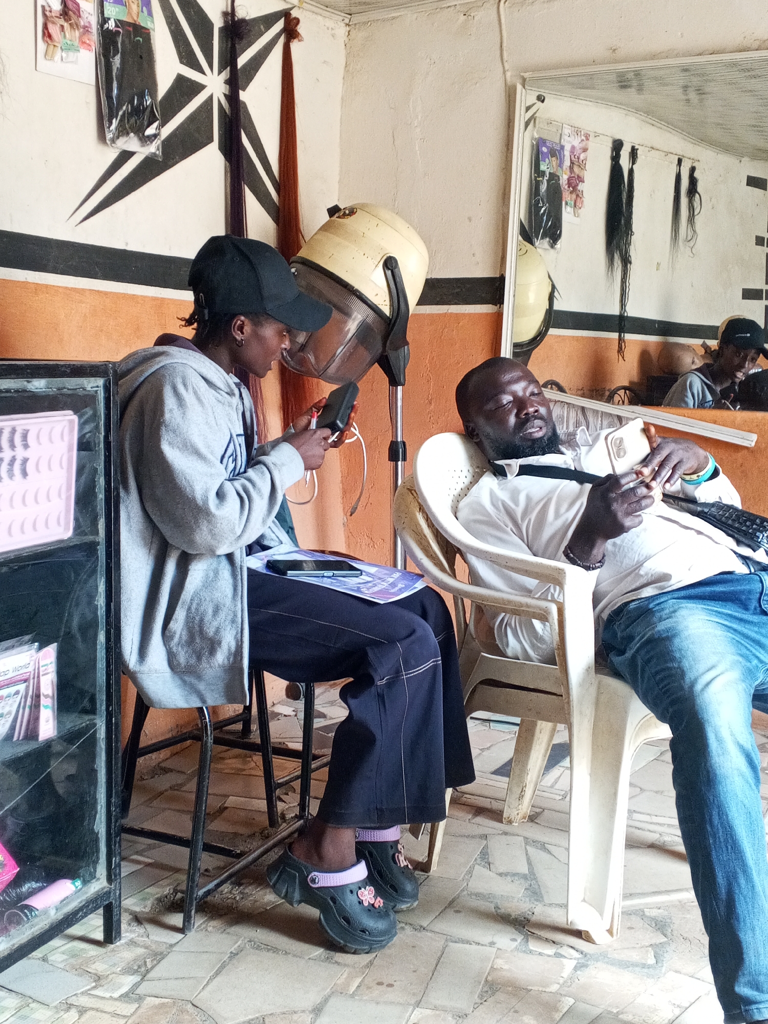 In a salon in an unspecified city, a person wearing a gray hoodie and black baseball cap sits under a hair dryer, manipulating an electronic device. They are seated on a stool next to a black display case filled with beauty products. Another person, wearing a white shirt and blue jeans, reclines in a white plastic chair, holding a smartphone. A mirror in the background reflects a partial view of the first person and salon decor, including hair extensions hanging on the wall. The floor has a tiled pattern, and the walls are painted with a horizontal stripe of orange.