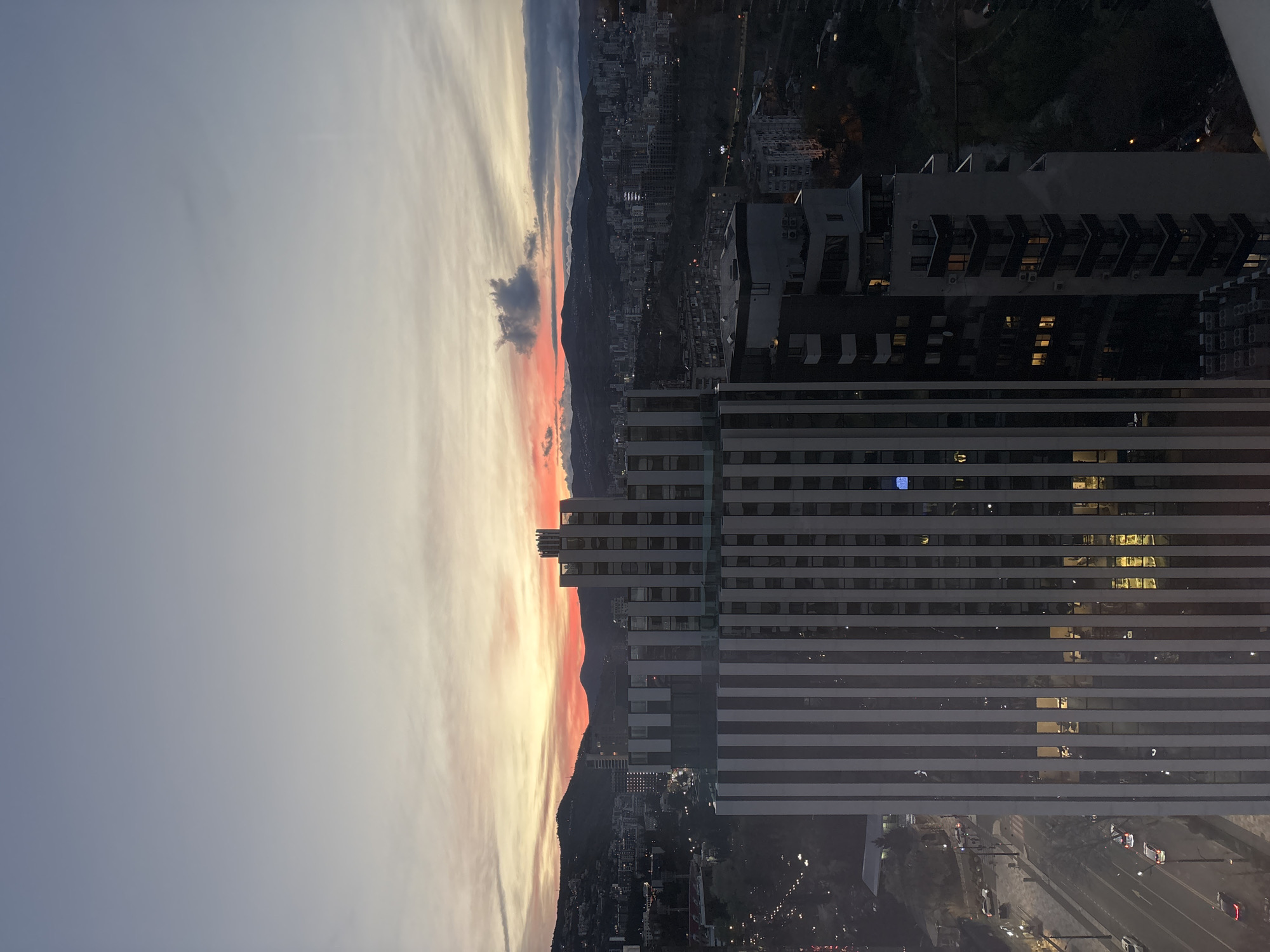 The image is an elevated view from a building overlooking a city, likely Tbilisi, Georgia, during a sunset or sunrise. The sky exhibits a gradient of colors ranging from light blue/grey to orange and red where it meets the horizon. Silhouetted mountains are visible along the horizon line. In the foreground, the view is dominated by a tall, modern building with horizontal stripes. At the bottom of the image, there is a glimpse of the street life with cars and streetlights. Other buildings and structures are visible behind the main building, with hints of urban activity.