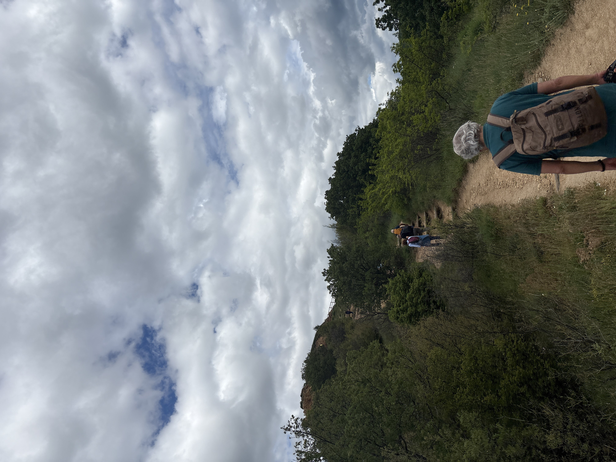 The image depicts a hiking trail in Stob, Bulgaria. A person wearing a green shirt and a brown backpack is walking uphill on the dirt path. Ahead of them, other hikers are visible further up the trail. The path is bordered by green trees and foliage. A rocky cliff face can be seen to the left. The sky is mostly cloudy with patches of blue.
