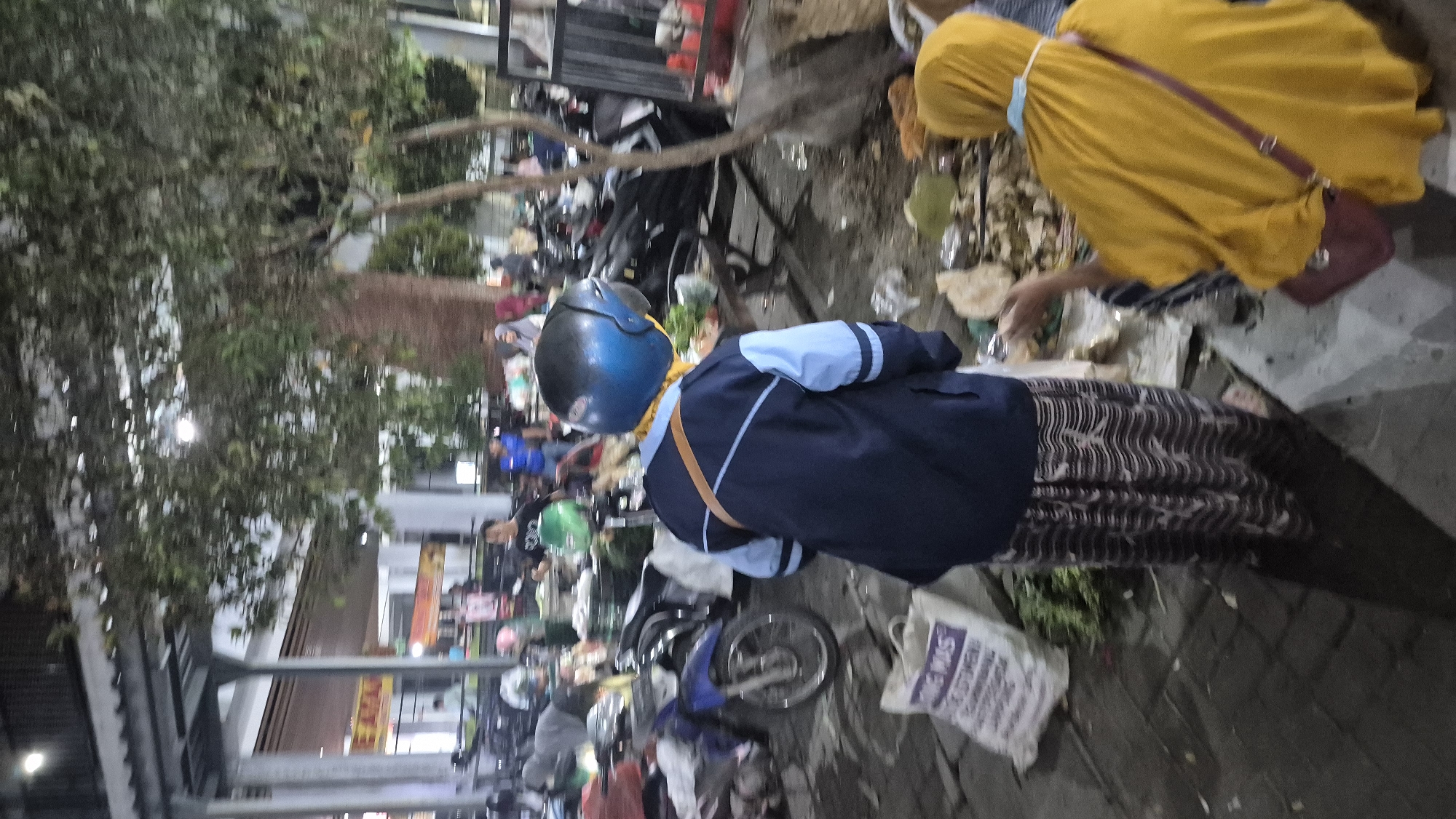 The image depicts an outdoor market scene at night in Bojonegoro, Indonesia. In the foreground, a person wearing a blue and light blue jacket, a helmet, and a patterned skirt is standing. To the right, another person in a yellow hijab is seated, facing away from the camera. Several motorcycles are parked in the background, along with market stalls and trees. The ground is paved with cobblestones. The overall impression is that of a busy, possibly bustling, night market environment.