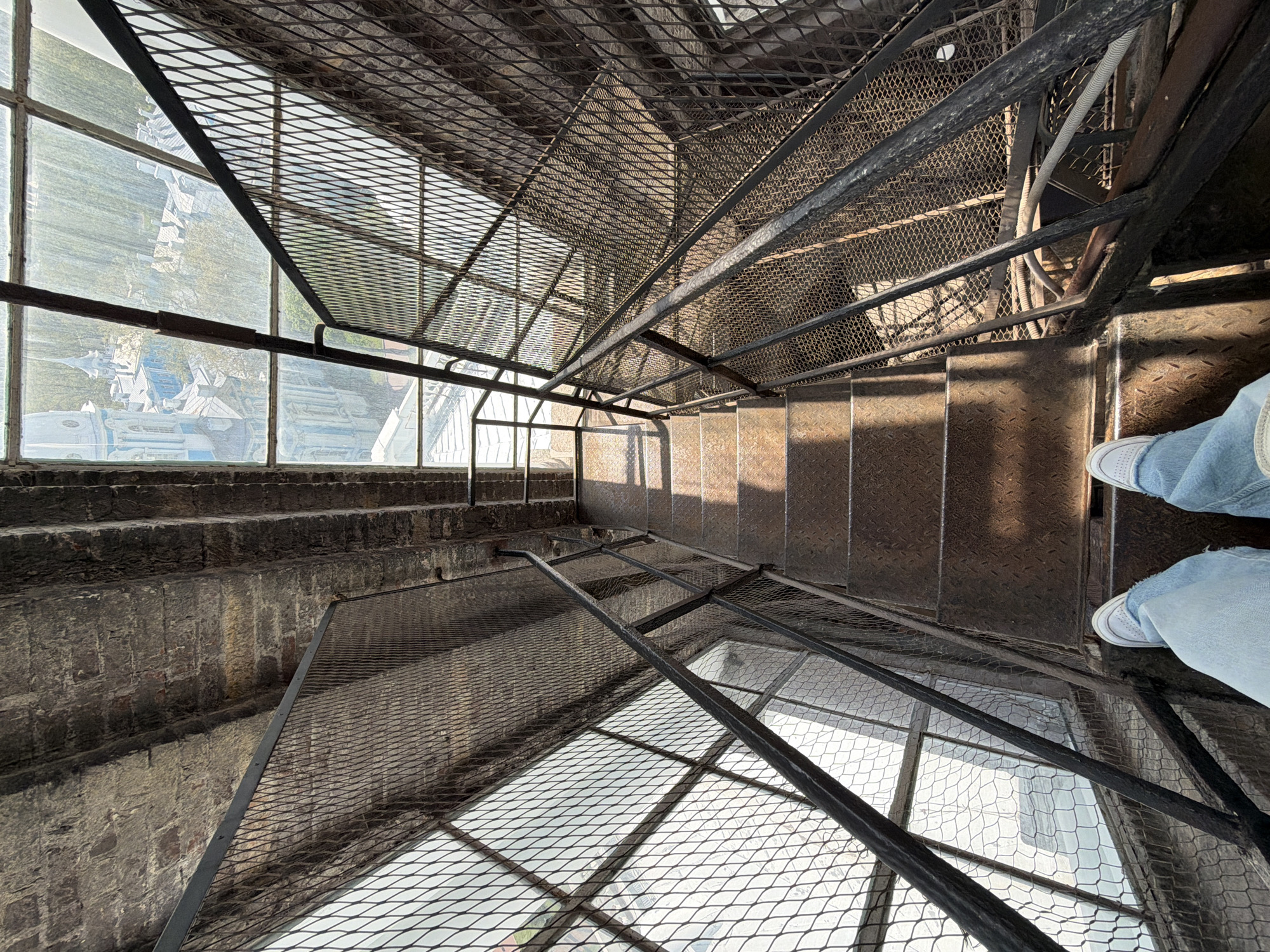 View looking down a metal staircase with diamond-patterned mesh railings. Exterior view visible through windows on the left side of the frame, reflected in the metal floor. The exterior view shows buildings and landscape. A pair of legs in light blue jeans and white sneakers are visible on the right side of the frame. The staircase is worn and shows signs of age. The location is Saint Petersburg, Russia.