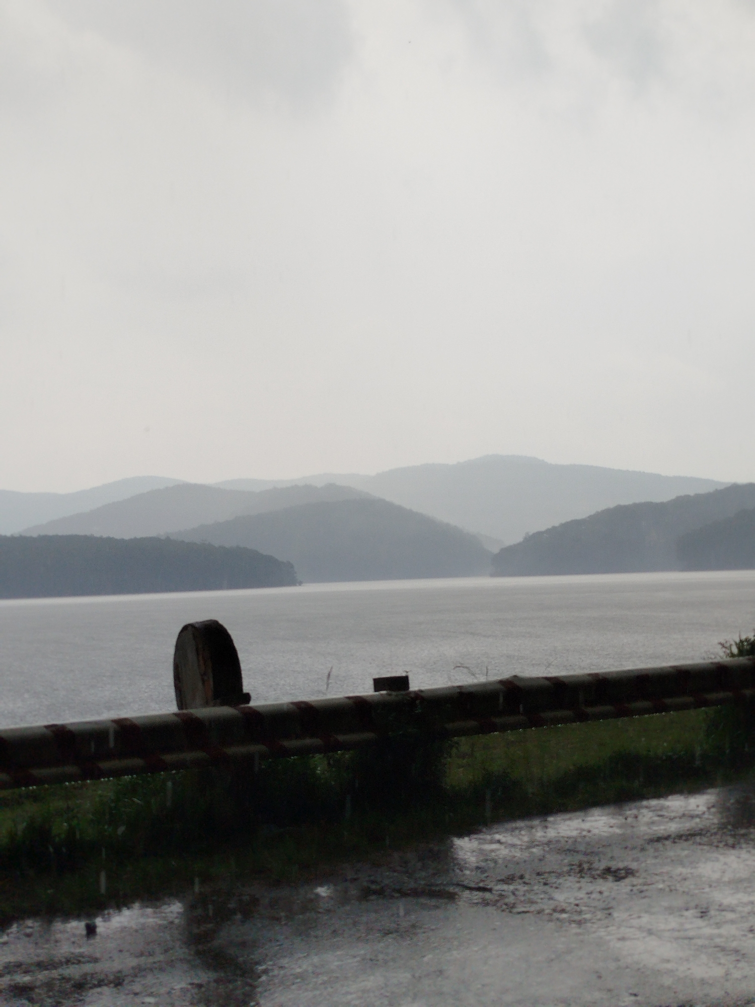 The photograph shows a landscape scene in Da Lat, Vietnam during a rainy day. In the mid-ground, there is a body of water, possibly a lake, with faint outlines of mountains in the background. The foreground has a metal guardrail with vegetation growing beneath it. The ground is wet, reflecting the heavy rain. The sky is overcast, contributing to the gray and somber tone of the image. No people are visible in the photograph.