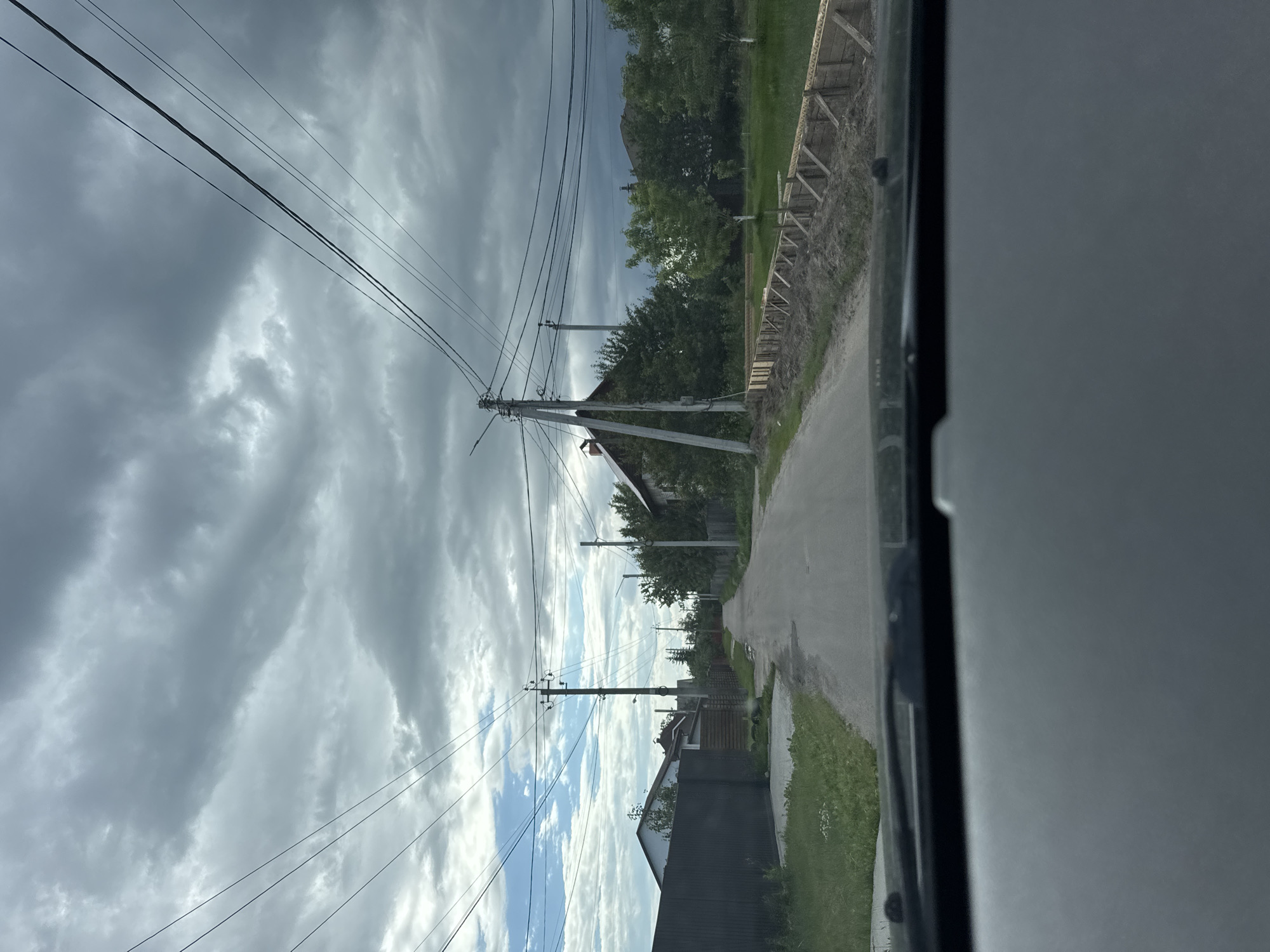 The image depicts a street scene in a rural or suburban area, viewed from inside a vehicle. The road is paved and appears to be a residential street, with houses and fences visible on both sides. A construction project is underway on the right side of the road, with wooden forms and dirt piled up, suggesting the beginnings of a foundation or retaining wall. Overhead, numerous power lines crisscross the sky, supported by utility poles. The sky is overcast with heavy, dark clouds, indicating potential for rain or stormy weather. The overall impression is one of a typical, somewhat dreary day in a developing or established residential area. There are no people visible in the image.