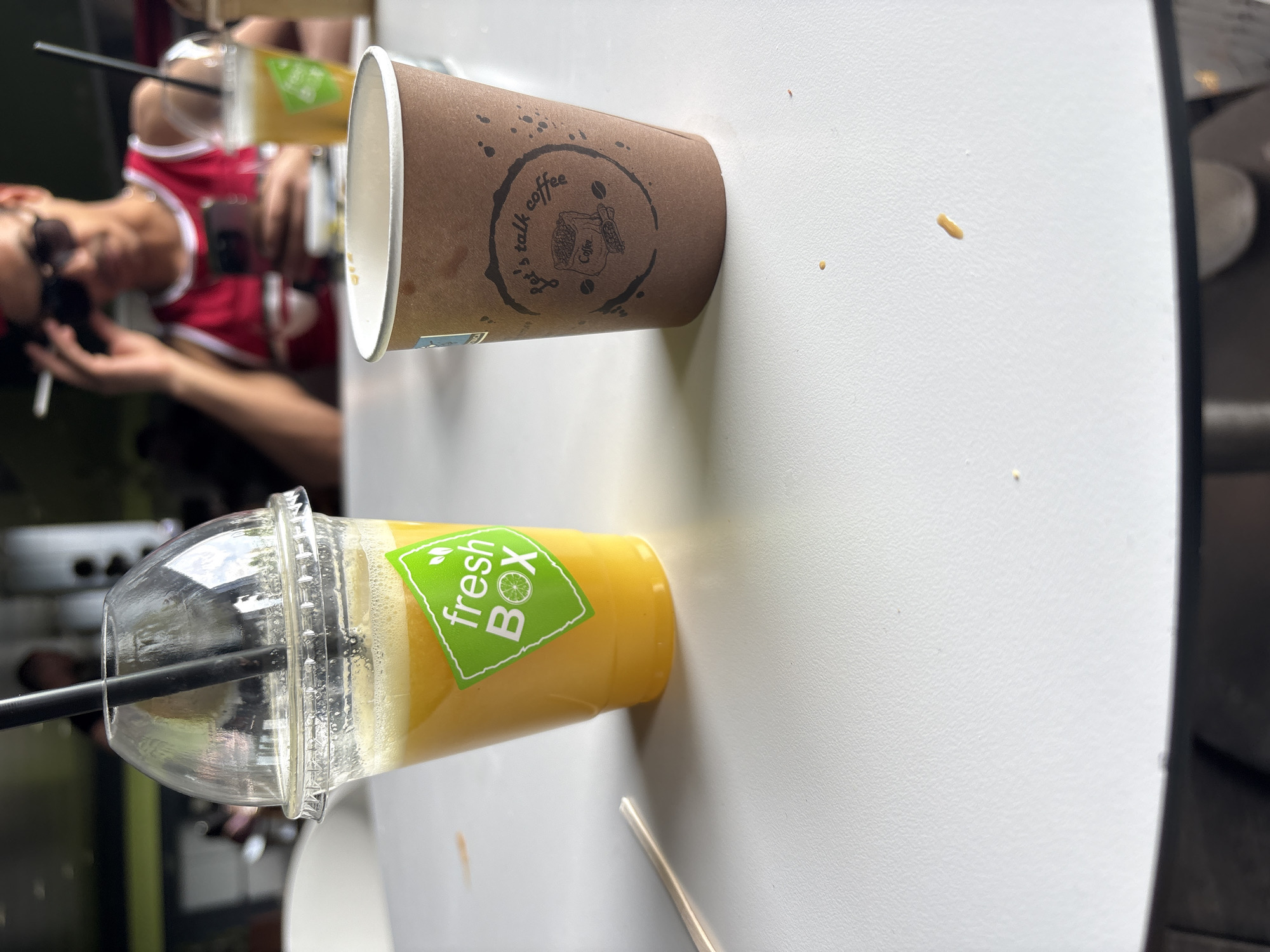 The image depicts a casual scene featuring two beverages on a white table, captured from an overhead perspective with the entire scene rotated ninety degrees clockwise. The objects thus appear sideways, as does a blurred person in the background. The setting suggests a daytime outing, possibly at an outdoor cafe in Plovdiv, Bulgaria.
On the left, a clear plastic cup, lying on its side, holds a vibrant orange or yellow beverage, likely juice or a smoothie, topped with a frothy layer. A black straw is inserted through its clear dome lid. A bright green label on the cup reads "fresh BOX," accompanied by a graphic of a lime or lemon slice.
Next to it, a brown paper coffee cup also lies on its side. Its exterior is adorned with a circular black logo featuring the text "Let's talk coffee" encircling an illustration of a coffee sack or bag and scattered coffee beans. The word "Coffee" is also visible within this central design.
The white table surface exhibits several small splatters or drips of a light brown or yellow liquid, suggesting minor spills from the drinks. A metallic spoon or stirring stick is partially visible near the bottom edge of the frame, close to the clear plastic cup.
In the blurred background, a person is visible, wearing a red and white athletic-style top and sunglasses perched on their head. They are holding what appears to be a lit cigarette or a similar object in their hand. The overall atmosphere conveys a sense of leisure and a relaxed break.