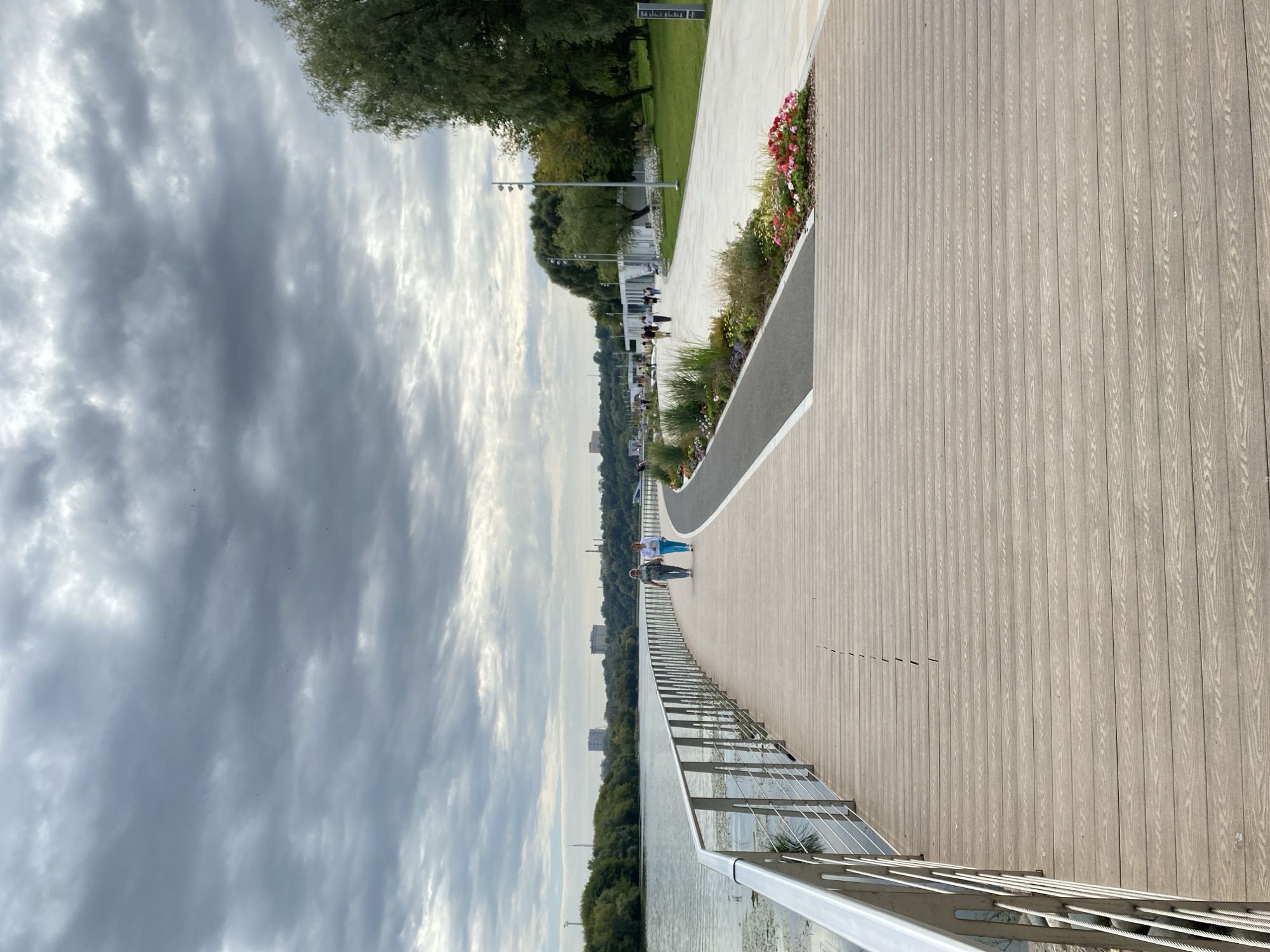 The image depicts a paved walkway along a body of water, likely a river or canal, in Moscow, Russia. The walkway is constructed of light-colored wooden planks and features a metal railing on the left side. A paved path runs parallel to the wooden walkway, separated by a strip of greenery with flowers.
In the middle ground, two women are walking on the paved path away from the viewer. They are dressed in casual clothing. Further along the path, other people are visible walking and congregating. Buildings and trees line the opposite bank of the water. The sky is overcast with dramatic gray clouds. The overall scene suggests a public park or recreational area, possibly the embankments of the Moskva River or a park like Sokolniki. The architecture of the distant buildings suggests an urban environment within Moscow.