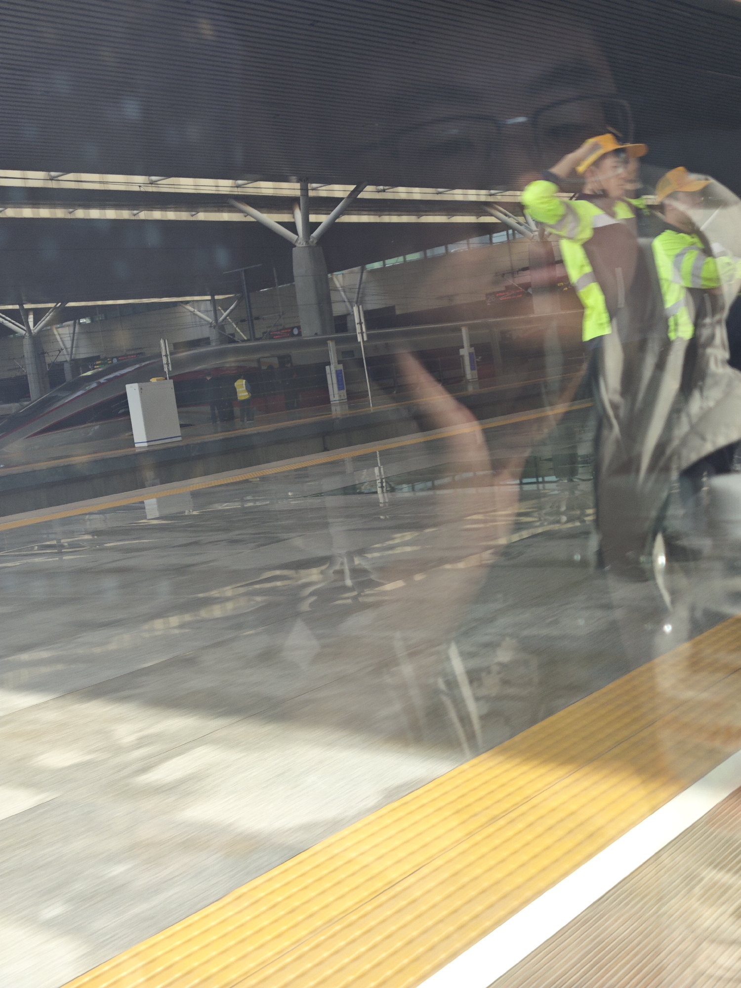 The image shows a train station with a modern, covered platform. A high-speed train is visible, parked at the platform. Two people wearing yellow hard hats and high-visibility vests are reflected in the window, appearing superimposed on the scene. There are yellow safety lines on the floor of the train station. The scene is observed through a window.