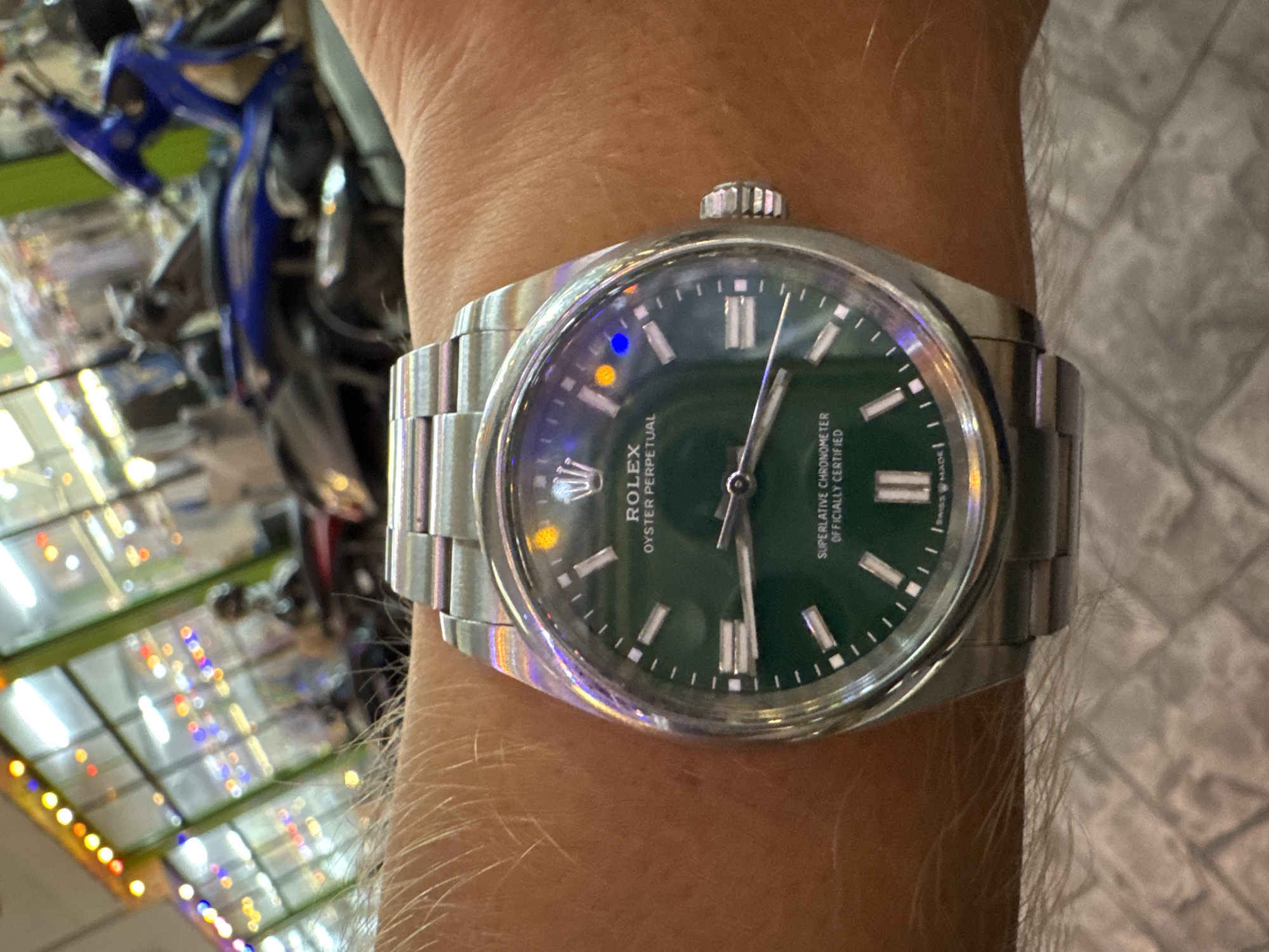 This is a close-up, eye-level, outdoor shot of a person's wrist wearing a silver Rolex Oyster Perpetual watch with a green dial. The watch has white hour markers and hands, with the Rolex crown logo at the 12 o'clock position. The text "ROLEX" and "OYSTER PERPETUAL" is visible on the dial, as well as "SUPERLATIVE CHRONOMETER OFFICIALLY CERTIFIED" and "SWISS MADE". The watch is reflecting light, with subtle rainbow colors visible on the bezel. The person's skin, covered in light hair, is visible around the watch. In the blurred background, a blue motorcycle can be seen, along with colorful lights suggesting a lively setting, possibly a market or entertainment area. The lighting suggests it is daytime, but the colorful lights could also indicate evening. The location is Karon, Thailand.