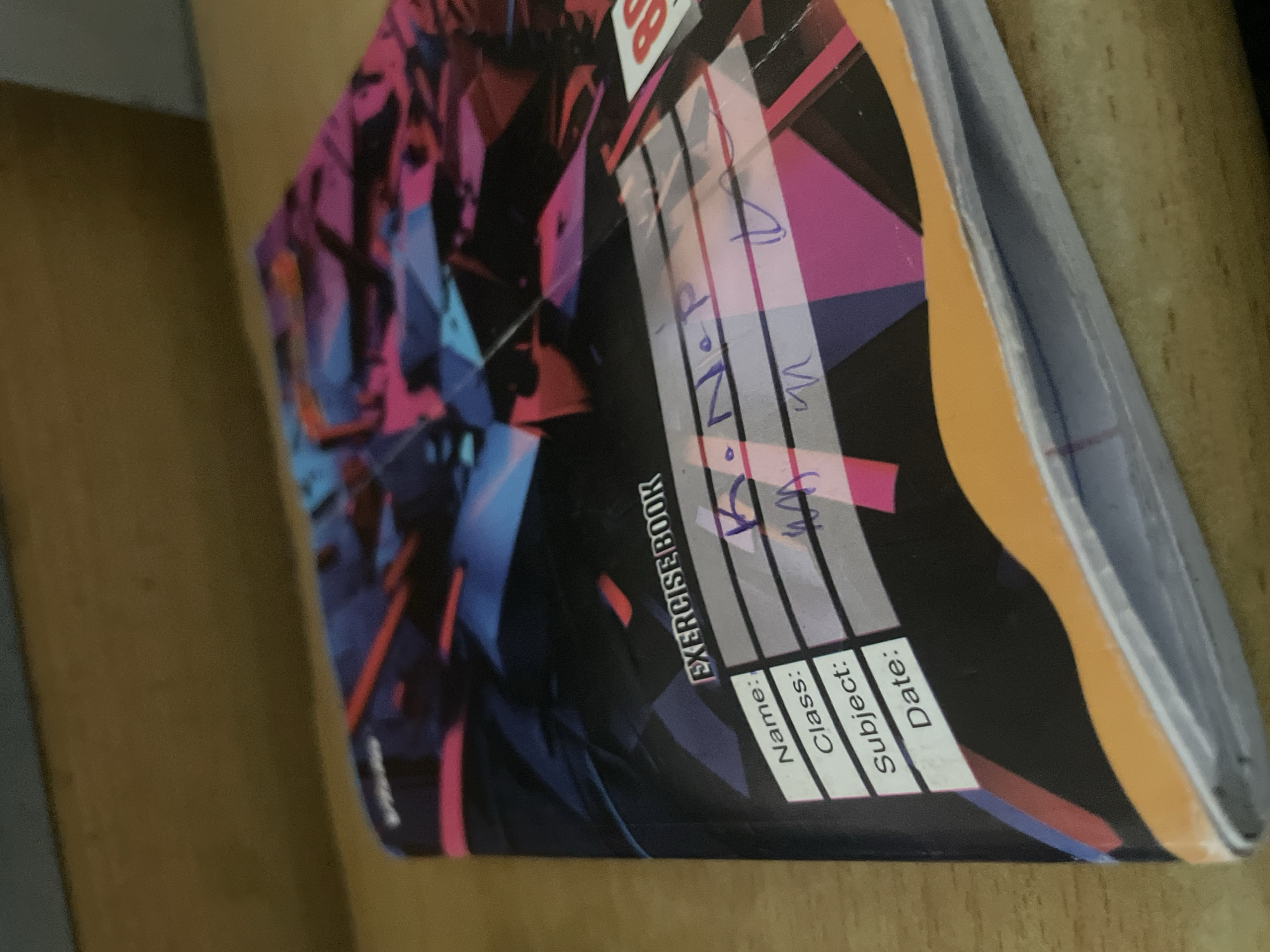 The image shows the cover of a notebook on a wooden surface. The notebook has a colorful, abstract design with geometric shapes in shades of blue, pink, and black. Printed on the cover are fields for "Name:", "Class:", "Subject:", and "Date:". Some of these fields have been filled in with blue ink. The "Name:" field appears to have "K. N. P" written in it. The "Class:" field has what looks like "m" written in it. The "Subject:" field has what looks like a handwritten "M" or stylized "W". The "Date:" field has a squiggle that may be a signature or date. The notebook is slightly angled, with its spine facing the viewer's left. The wooden surface appears to be a desk or shelf. The lighting suggests an indoor environment. The image does not contain any information that allows for the identification of a specific location in the city of Ihiala, Nigeria.