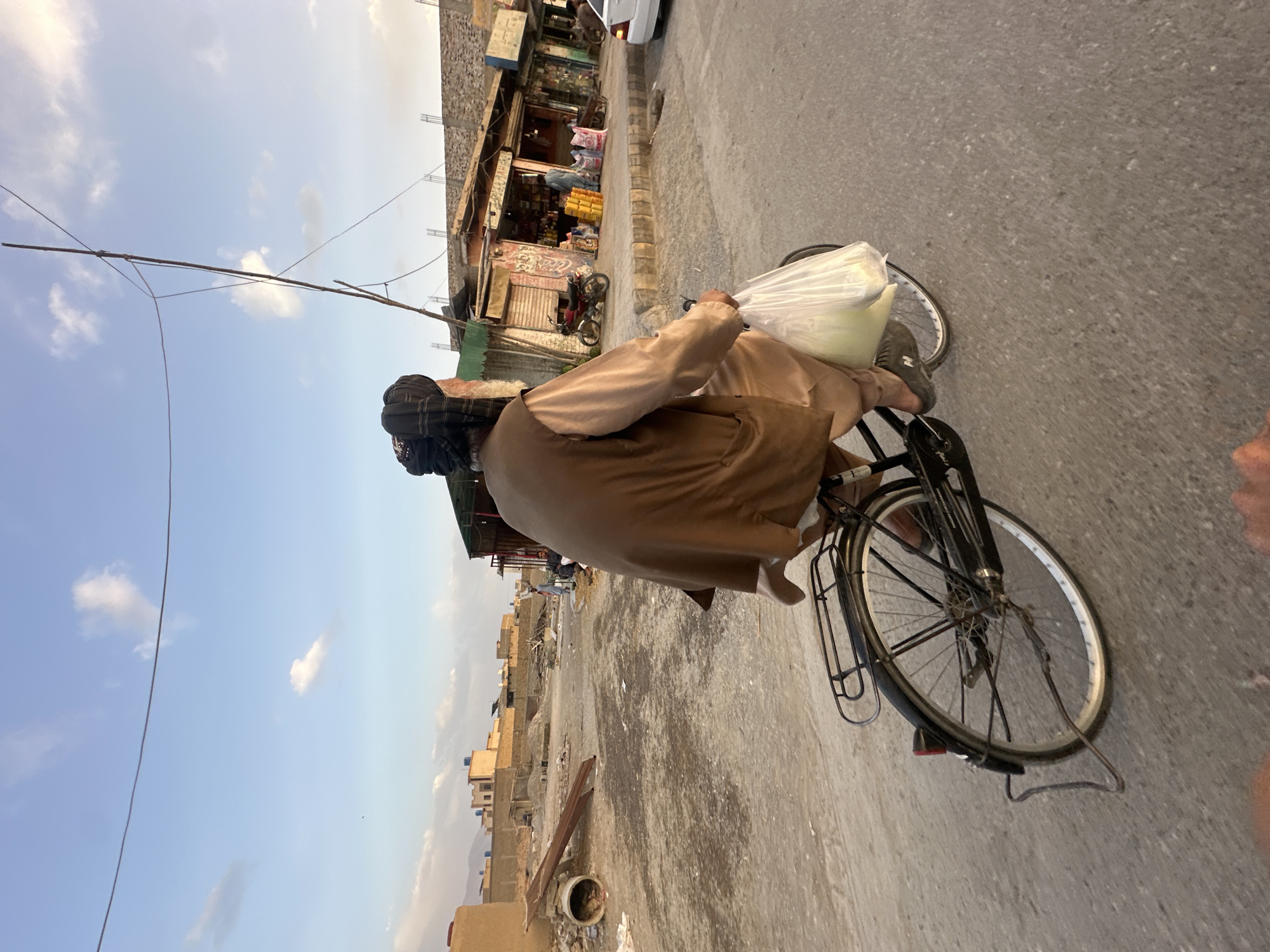 A man wearing a head covering rides a bicycle on a street in Quetta City Tehsil, Pakistan. He carries a white plastic bag on the handlebars. Buildings line the street, and the sky is partly cloudy. There is a power line above the man's head.