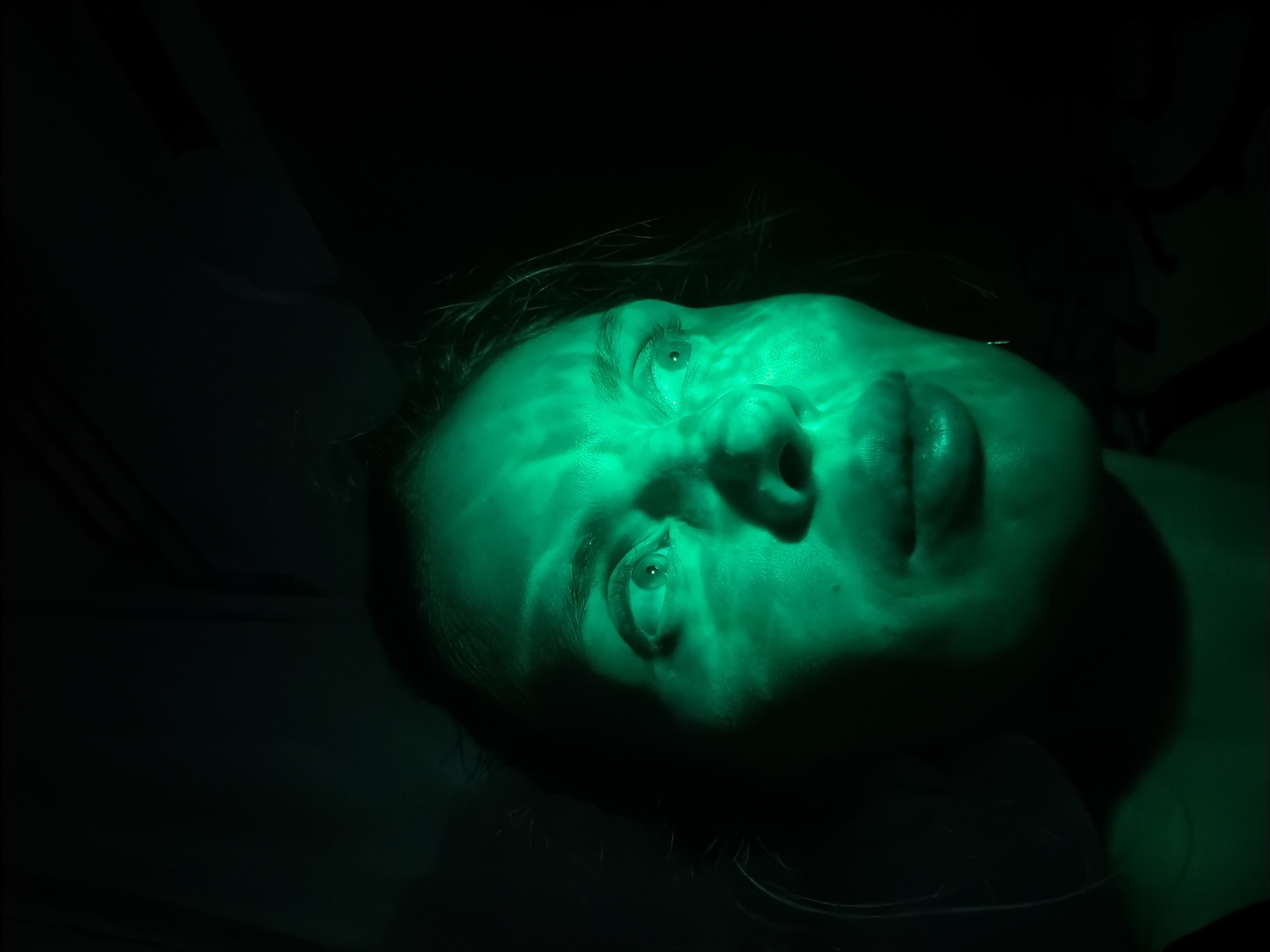 This striking image presents a close-up of a person's face, illuminated by a vivid, emerald green light against an almost entirely dark background. The subject, appearing to be female, is positioned with her head tilted, and her wide-open eyes gaze upwards and slightly to the right, conveying an intense or contemplative expression. The strong green light casts dramatic shadows across the contours of her face, highlighting her nose, lips, and the area around her eyes.
Distinct wavy patterns of light are visible across the illuminated skin, particularly on the forehead and cheeks, suggesting reflections from a rippling surface, perhaps water, or a specialized light projection. Strands of dark hair are visible around the periphery of her face, blending into the surrounding darkness. The environment is otherwise featureless, emphasizing the dramatic interplay of light and shadow on the subject. The overall mood is surreal and mysterious, with the unusual green illumination giving the scene an otherworldly quality. No specific objects or text are visible, aside from the person and the distinctive lighting. Given the location context, the scene is set in Budva, Montenegro, though there are no explicit visual cues within the image itself to confirm this.