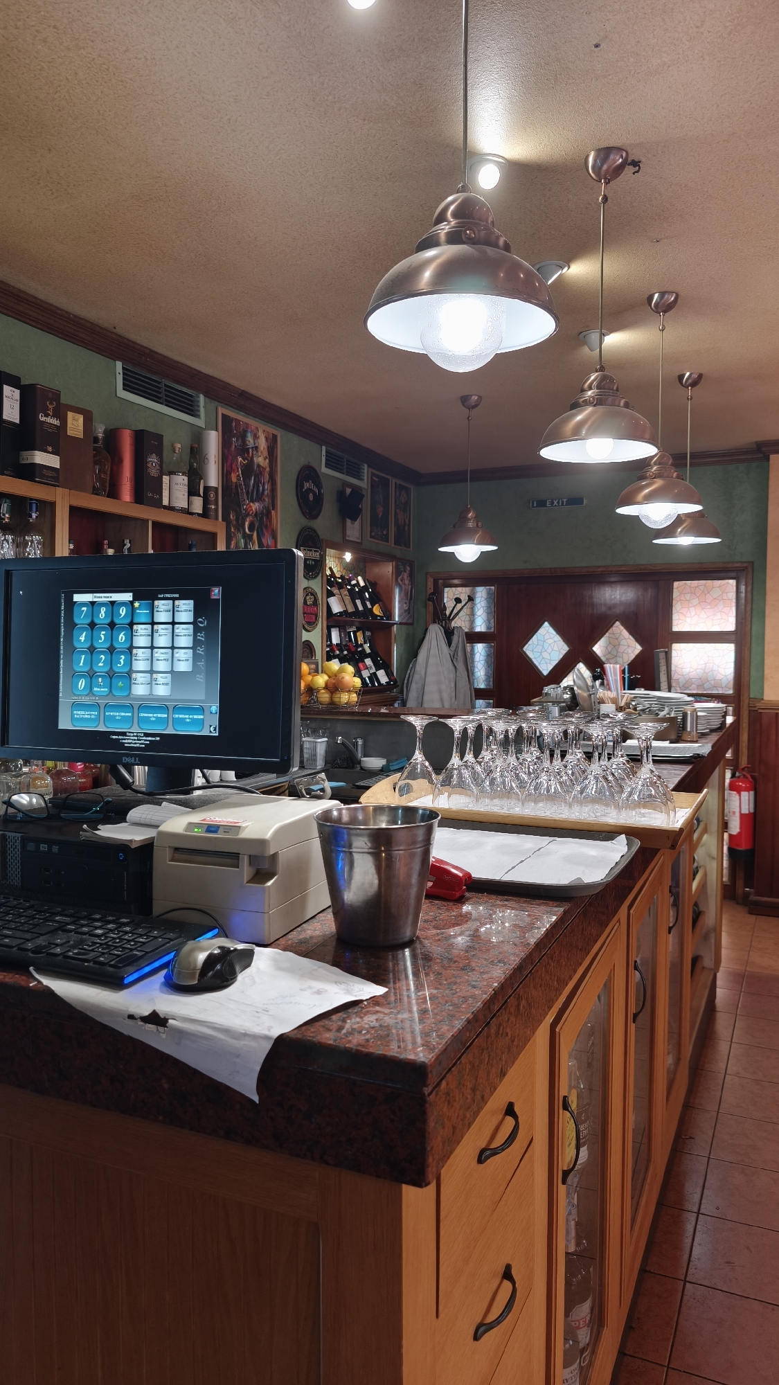 The image shows the interior of a bar or restaurant, likely in Pomorie, Bulgaria. The scene is dominated by a counter area with a computer monitor displaying a point-of-sale system, a keyboard, a mouse, and a receipt printer. To the right of the computer, there is a metal bucket, a red object, and a tray of wine glasses. Behind the counter, shelves are stocked with bottles of alcohol and wine. The walls are decorated with framed pictures and posters. The ceiling features multiple pendant lights, and an "EXIT" sign is visible in the background. The overall impression is that of a functional service area within a hospitality establishment.