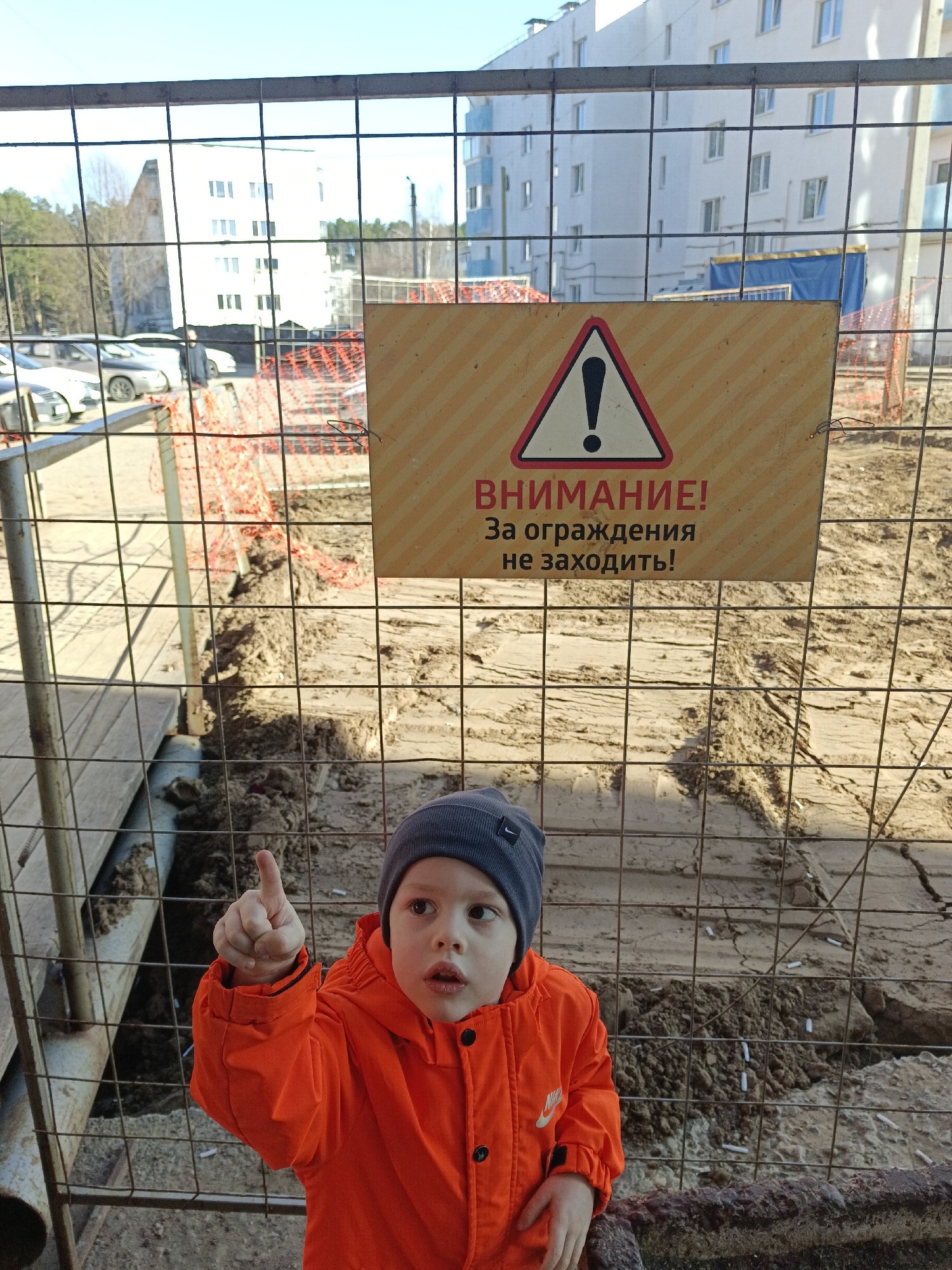 A young boy wearing a gray beanie and an orange jacket stands in front of a wire fence in Chaikovsky, Russia. He points upwards with his right index finger. Behind the fence, there is a construction site with a sign in Russian that reads "ВНИМАНИЕ! За ограждения не заходить!" ("ATTENTION! Do not enter the fence!"). The ground inside the fence is dirt and sand, with visible trenches. In the background, there are multi-story white buildings, trees, and parked cars. Orange safety fencing is also visible. The scene is lit by bright sunlight.