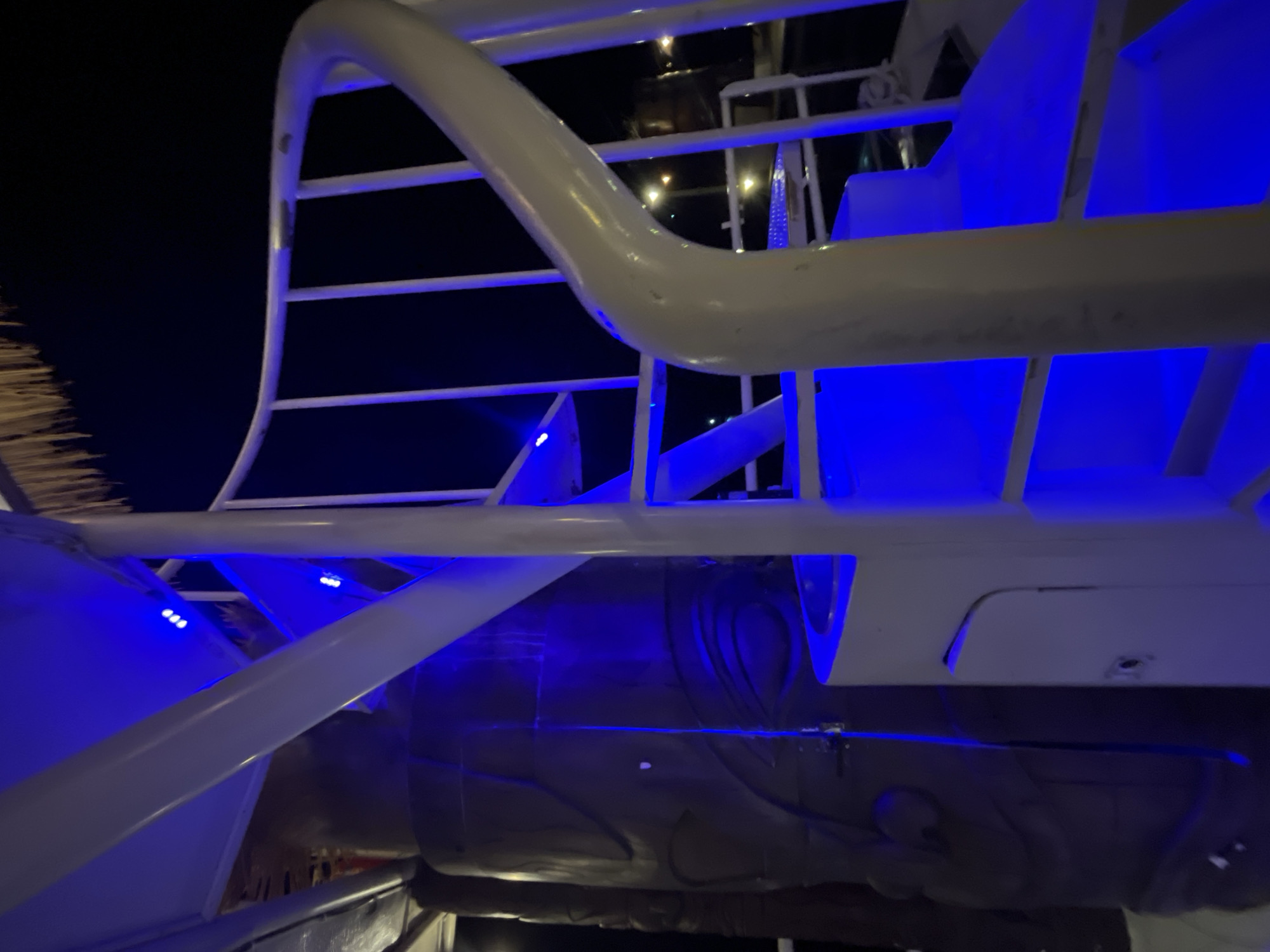 A low-angle, medium shot captures a spiral staircase illuminated by blue LED lights at night, likely on a boat or resort in Karimunjawa, Indonesia. The staircase, made of white metal, curves upwards and to the right, with vertical railings. To the left of the staircase is a large, dark brown tiki statue with carved facial features, its texture appearing rough and weathered. The background is dark, suggesting it's nighttime, with faint lights visible in the distance. The scene has a festive and slightly mysterious atmosphere due to the blue lighting.