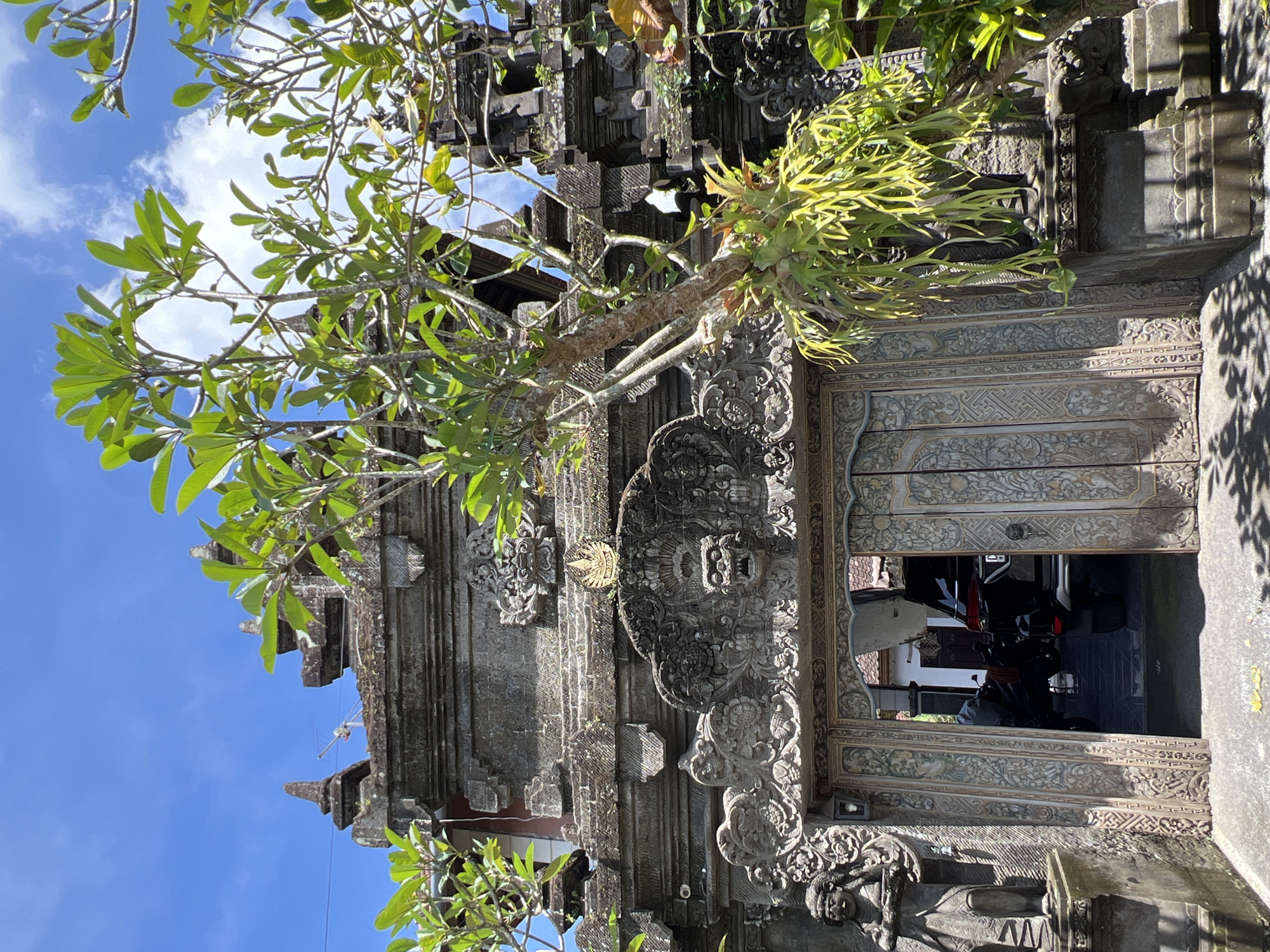 The image captures a detailed, ornate stone entrance in Peliatan, Indonesia. A leafy tree with green foliage frames the left side of the structure against a blue sky with scattered clouds. The stone entrance is adorned with intricate carvings and sculptures, displaying a rich, aged texture. Inside the doorway, a glimpse of a courtyard reveals a parked black vehicle. The scene is well-lit, accentuating the architectural details and the interplay of light and shadow.