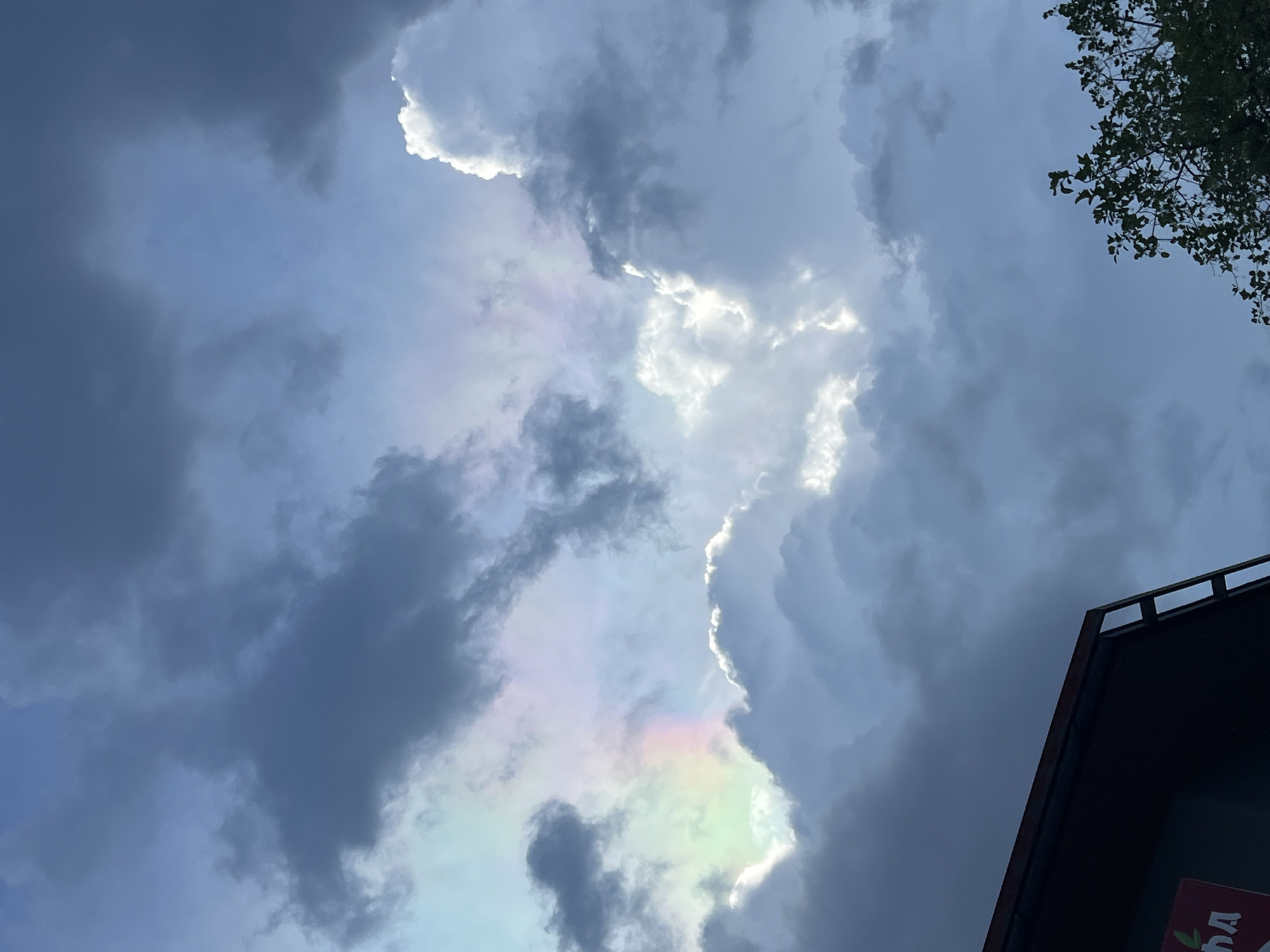 A low-angle shot of clouds is seen in the sky. A patch of iridescence is visible through the clouds, displaying faint colors of the rainbow. Overlapping layers of storm clouds are situated beside the iridescent patch. In the top-right corner of the frame, some tree branches are visible. In the bottom-right corner, the edge of a building roof is apparent. Based on the metadata, the location is Baan Bo Phut, Thailand.