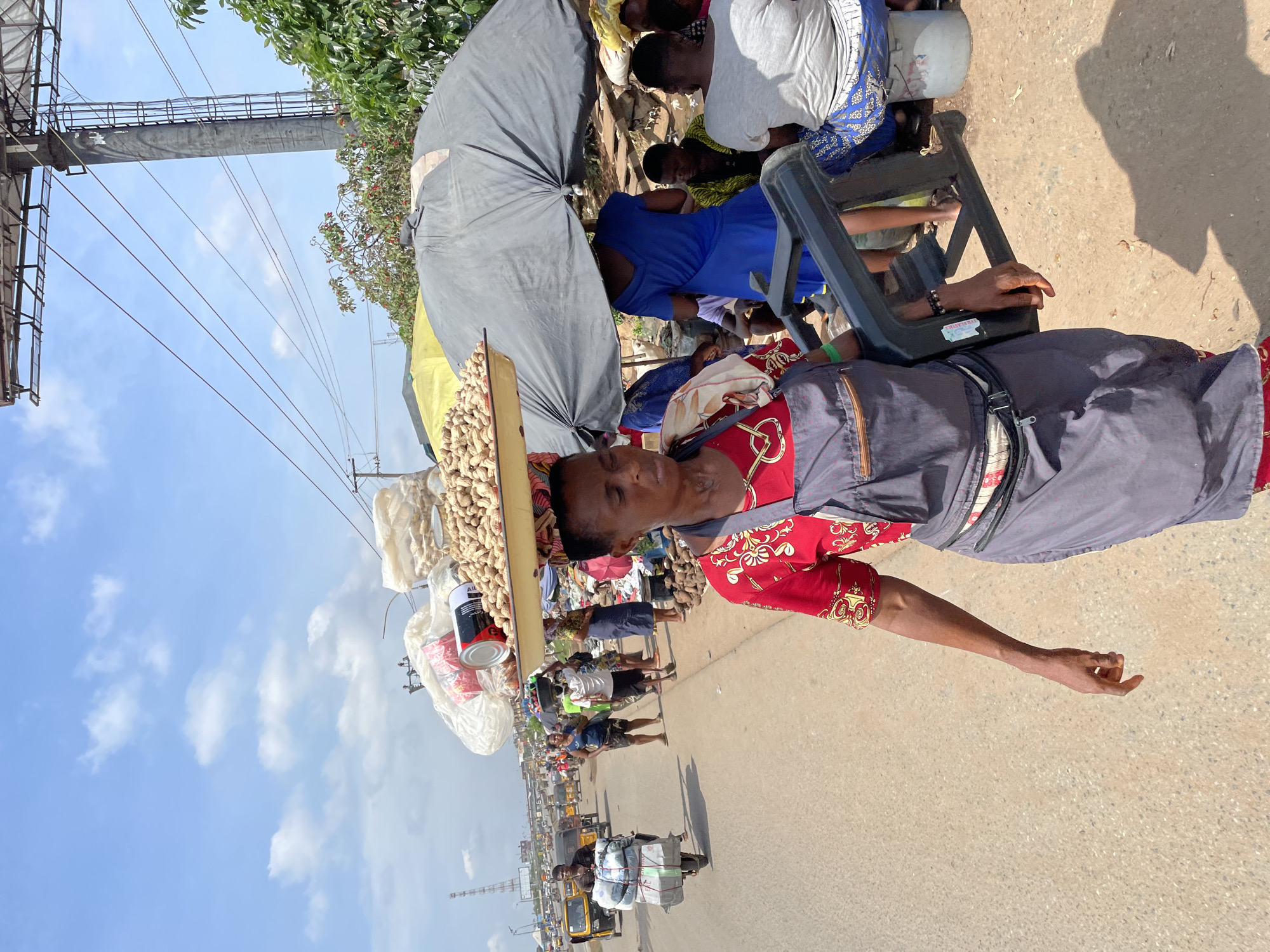 A full-shot medium-distance view shows a street scene in Onitsha, Nigeria, on a sunny day. A woman in the foreground walks towards the left, carrying a large tray of peanuts balanced on her head. She wears a red shirt with gold and black patterns, a grey apron over dark pants, and a fanny pack around her waist. She is also carrying a black plastic chair in her right arm. Behind her, to the right, a large black umbrella shades several people sitting on stools or the ground. In the background, more people and market stalls are visible, with traffic including motorcycles and a tricycle. The road is paved, and power lines crisscross the sky above.