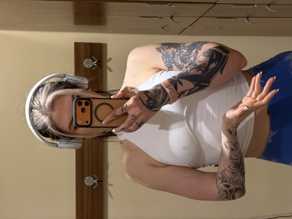 A female-presenting individual is depicted taking a mirror selfie in what appears to be a changing room or gym facility in Budva, Montenegro. The subject has dark hair with prominent white/blonde streaks at the front and wears white over-ear headphones. She is dressed in a white ribbed halter-neck crop top and blue patterned leggings. Her face is largely obscured by a smartphone, which is held in her right hand and features an orange case with a black circular magnet attachment and a triple-lens camera module. Tattoos are visible on both arms, including abstract designs on her left forearm and detailed figurative art on her right upper arm and forearm, which includes faces and a cat-like creature. A light pink fitness tracker or strap is present on her right bicep. Small tattoos are also visible on her fingers and hands. The background consists of a light beige wall with a wooden rail featuring two metal double-pronged hooks on the left. To the right, a vertical stack of light-colored lockers is visible.
