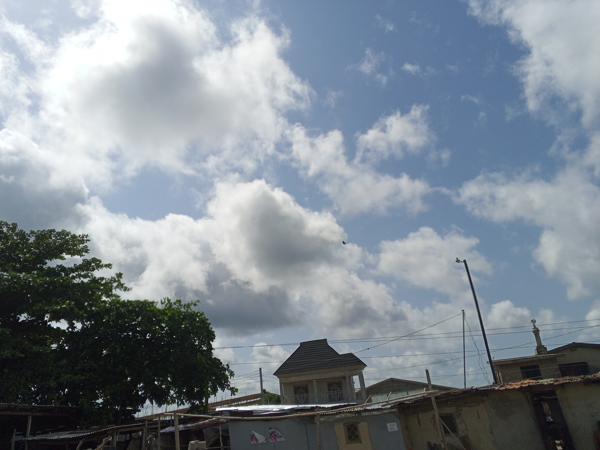 The image presents an urban scene, possibly in the city of Badagry, Nigeria. The sky is partly cloudy with a mix of white and gray clouds against a light blue backdrop. In the foreground, a large green tree stands to the left. Several buildings of varying sizes and conditions are visible. Some appear to be temporary structures, while others have more permanent construction, one with a second-story balcony. A thin street light is standing near the center of the image. Electrical wires stretch across the sky. A small religious sculpture is visible above the building in the middle right, likely a mosque. A single bird can be seen flying in the sky above the building with the second-story balcony.