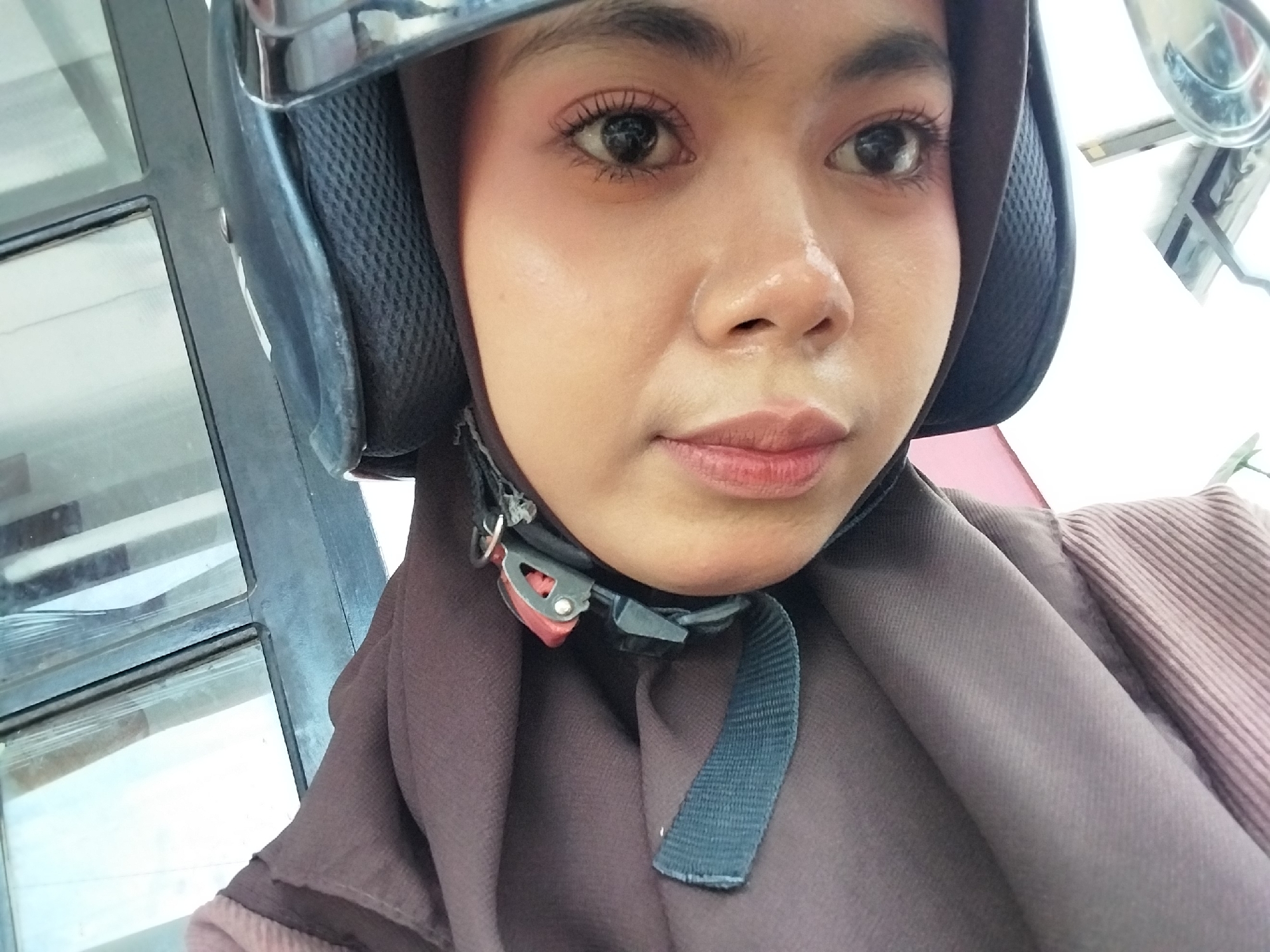 A woman is taking a selfie while wearing a helmet and a hijab. The helmet is black and has a silver trim. The hijab is a dark brown color. The woman is wearing a light pink shirt. Her makeup includes mascara and lipstick. She is looking at the camera. The background appears to be outdoors, with a window and some blurry objects visible. The location is stated to be in the city of Tibojong, Indonesia.