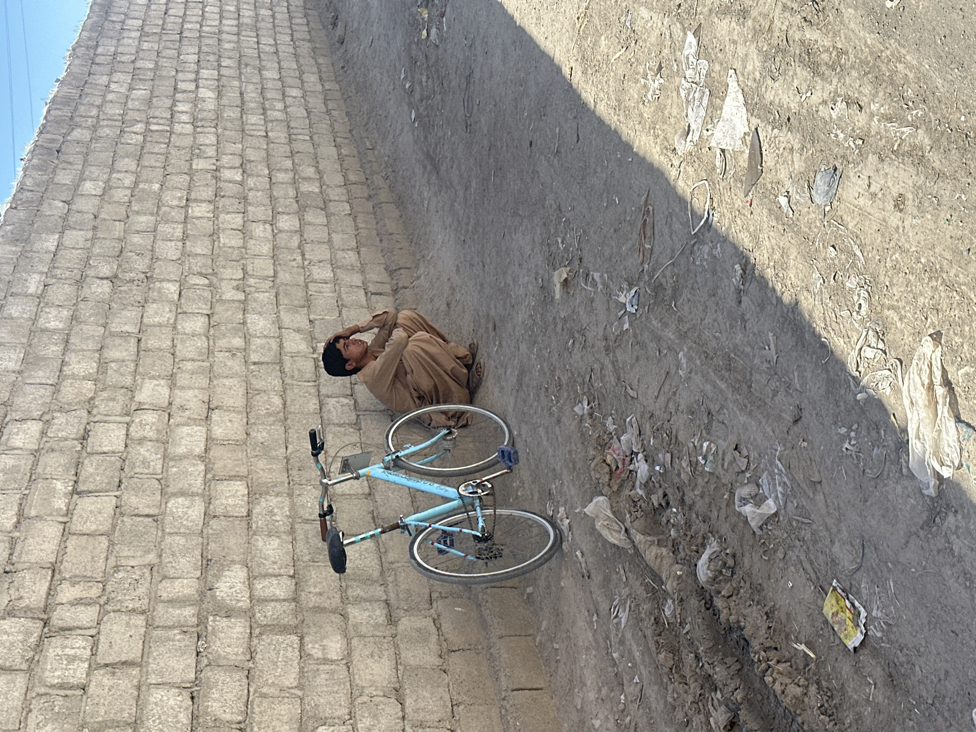 A person is sitting next to a light blue bicycle beside a paved block wall in Quetta City Tehsil, Pakistan. The person wears tan clothing and has their head in their hands. Trash and debris litter the ground near the edge of the wall and on the dirt road. Shadow falls across the road.