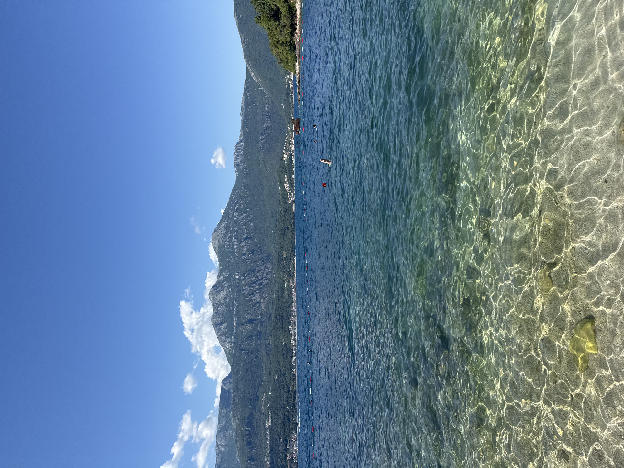 The image depicts a panoramic view of a bay or a large body of water on a clear, sunny day, with a majestic mountain range in the background and a forested coastline on the right, in the city of Unknown, Unknown. The sky above is a vibrant blue with scattered white cumulus clouds, particularly visible above the mountains on the left. The mountains are rugged, steep, and covered in dense green vegetation, with visible rock faces. Along the base of the mountains, a coastal settlement with numerous buildings and houses stretches across the middle distance.
In the foreground, the water is exceptionally clear, revealing the sandy or pebbly seabed with distinctive patterns of sunlight refracting through the water's surface. As the water extends towards the mid-ground, it transitions to a deeper blue. A discernible line of red and white cylindrical buoys stretches across the mid-distance, likely marking a swimming area or boundary. Within this water, a single person is visible, partially submerged with both arms raised out of the water, suggesting a swimming motion like a backstroke or a dive. Further back and slightly to the right of the person, a reddish-brown, partially submerged structure, resembling a derelict boat or shipwreck, is visible. On the far right, a densely forested hill or landmass slopes down to meet the water. The scene is illuminated by bright daylight, casting strong reflections on the water's surface.