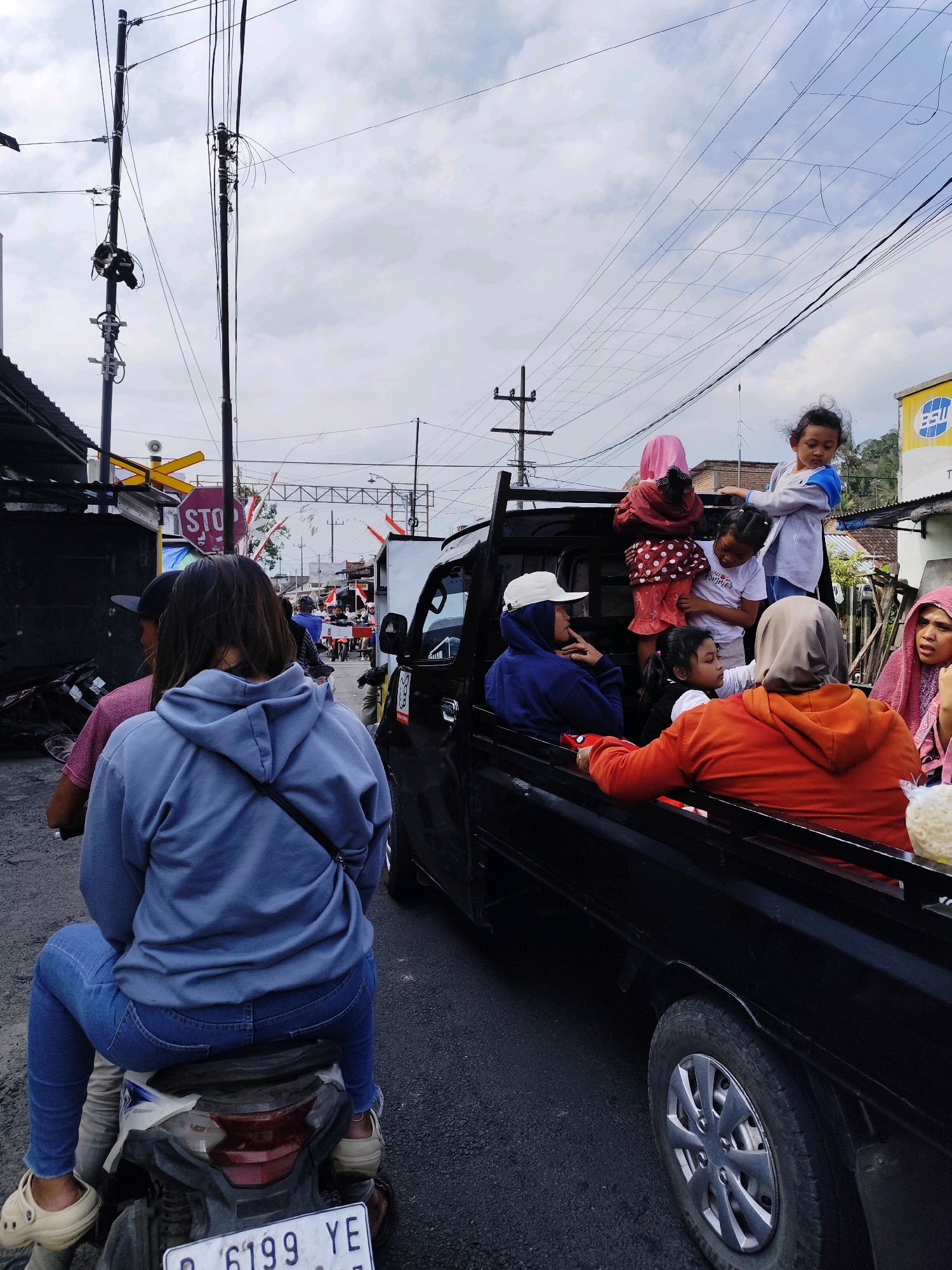 In the city of Jambewangi, Indonesia, a scene unfolds on a cloudy day on a paved road. A woman wearing a blue hoodie and jeans is seated on a motorcycle, facing away from the camera. She is positioned on the left side of the frame, with her back to the viewer. To her right, a black pickup truck is present, carrying several individuals.
Inside the truck, a group of people, including adults and children, are being transported. One woman in the truck is wearing an orange hoodie and a grey headscarf. Another woman, to her right, wears a pink hijab. Children are also visible, with one young girl in a grey long-sleeved shirt and another in a polka-dot outfit. A person wearing a blue hoodie and a white cap is seated inside the truck, facing left.
The environment includes utility poles with numerous wires overhead, a building with a dark facade to the left of the frame, and a yellow and red "STOP" sign with a barrier gate in the background. The road surface appears worn. The overall impression is of a busy street scene, likely during daytime.