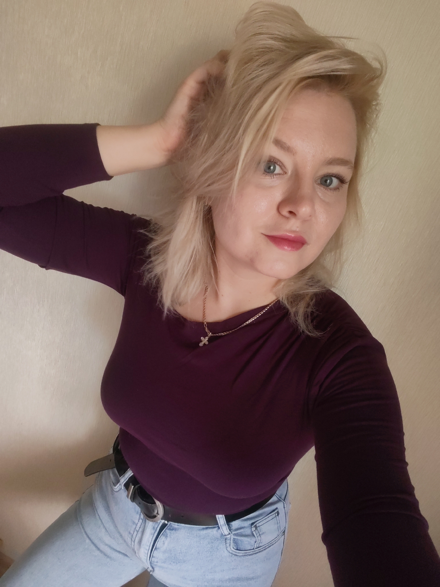 A young Caucasian woman with blonde hair and blue eyes, wearing a maroon long-sleeved shirt and light blue jeans, is captured in a selfie. She has her right hand raised, touching her hair, and is looking directly at the camera. She is wearing a necklace with a small cross pendant and a black belt. The background appears to be an indoor wall with a textured surface. The lighting is soft and diffused. There is no discernible information about the location being in Izhevsk, Russia.