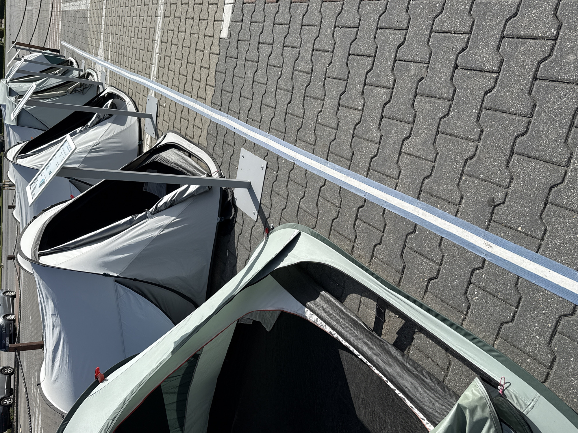 The image depicts a row of grey and light green pop-up tents displayed in an outdoor setting, likely a parking lot or an open area paved with interlocking grey concrete pavers. A blue and white striped line runs along the pavement in front of the tents. In the foreground, a light green tent is open, revealing its dark interior and carbon fiber poles. Behind it, a series of grey tents are set up, some with information stands in front of them. The background shows two parked cars and a dark chain-link fence. The scene appears to be in Bielany Wrocławskie, Poland, as indicated by the presence of Polish license plates on the cars.