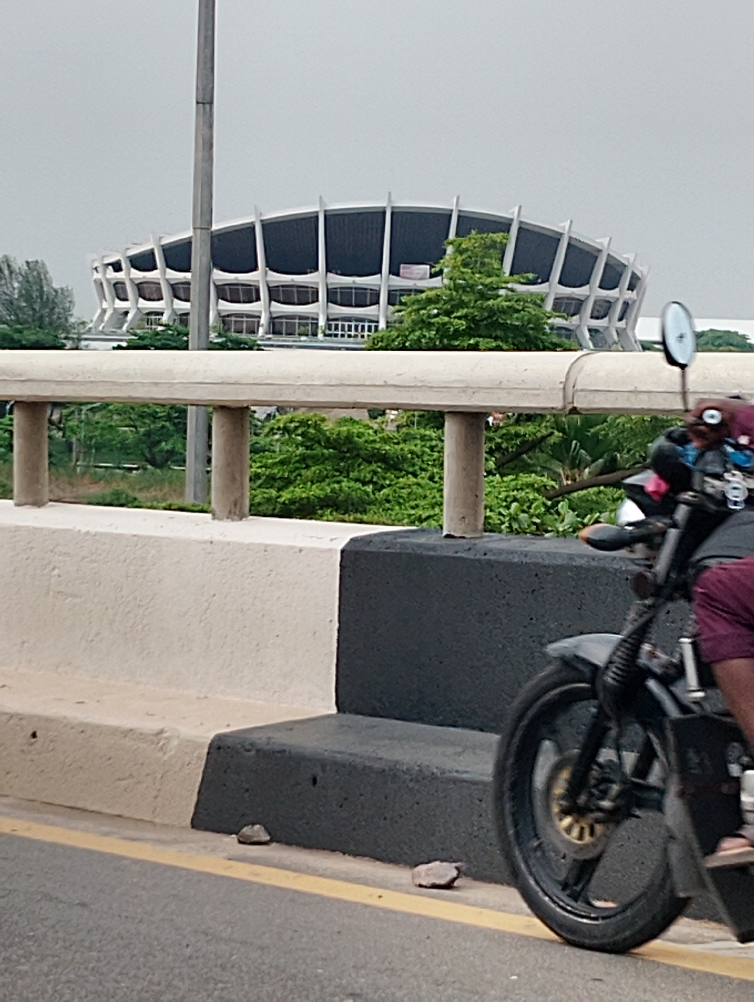 The image captures a view from a bridge in Alagbede, Nigeria. In the background, the distinctive white and dark facade of a large, curved building, likely a stadium or convention center, dominates the skyline. Lush green trees surround the structure, softening its imposing presence. In the foreground, a motorcycle, partially obscuring the view, is seen from the side, with its rider's legs in maroon shorts visible. The motorcycle has a black frame and wheel, with gold accents on the hub. A prominent concrete barrier, painted white and black, lines the edge of the bridge, separating the road from the embankment. A yellow line marks the lane on the asphalt road. The sky is overcast, suggesting a cloudy day. The overall scene depicts a moment of transit on a bridge, with a prominent landmark in the background.