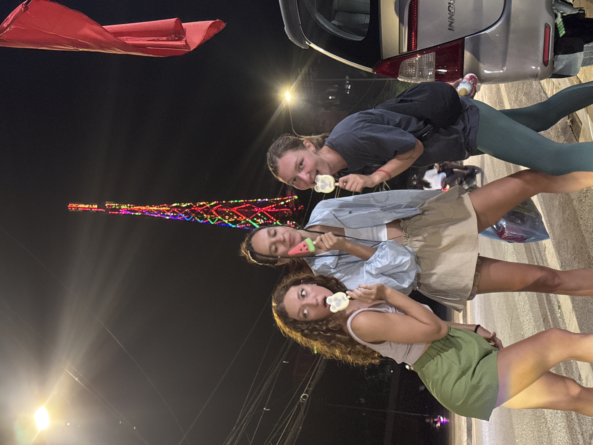 Three young women stand in a row at night in Yên Minh Commune, Vietnam, each holding a uniquely shaped lollipop. The backdrop features a decorated Christmas tree-like structure, illuminated streetlights, and overhead power lines. A red flag flutters in the upper left of the image. The women wear casual clothing: the bottom woman wears a green skirt and light pink top, the middle woman a beige skirt and blue denim shirt, and the top woman green leggings and a dark t-shirt. The rear of a silver vehicle is visible in the upper right corner.