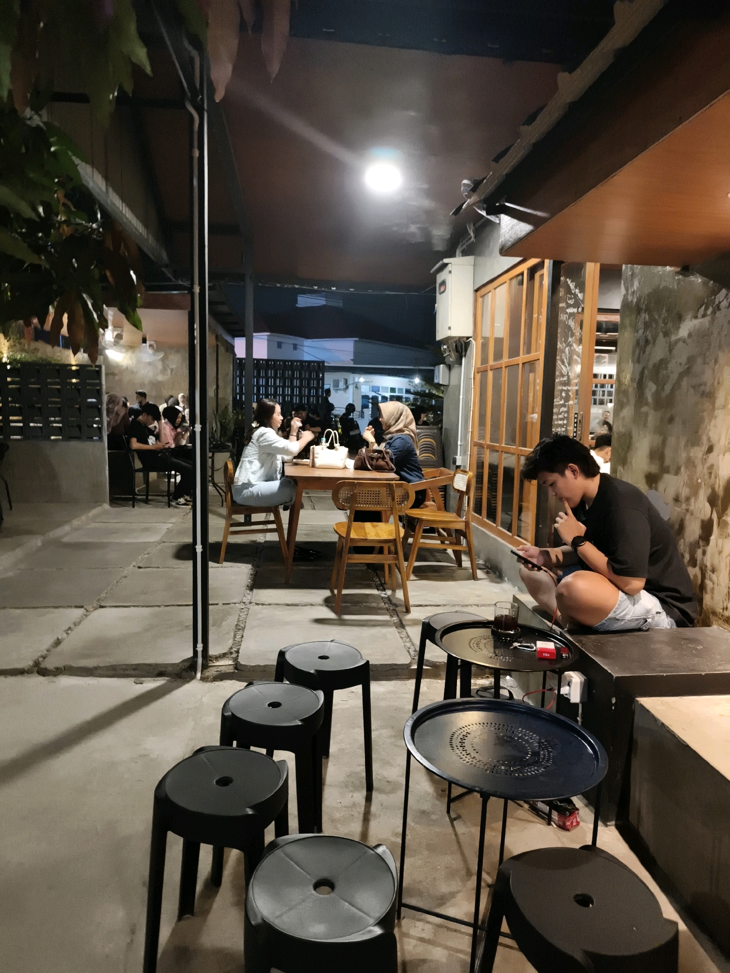The image depicts an outdoor seating area of a cafe at night, likely located in Ketapang, Indonesia. Several individuals are present, with two women seated at a table in the mid-ground, engaged in conversation. A man in the foreground is seated on a low bench, looking at his phone. The cafe features a concrete floor with a patterned design, black stools, and minimalist black tables. Wooden chairs are also visible around the tables. The cafe has an open-air roof supported by metal beams, with warm lighting illuminating the space. The background shows a building with large windows and an adjacent wall with a textured finish.