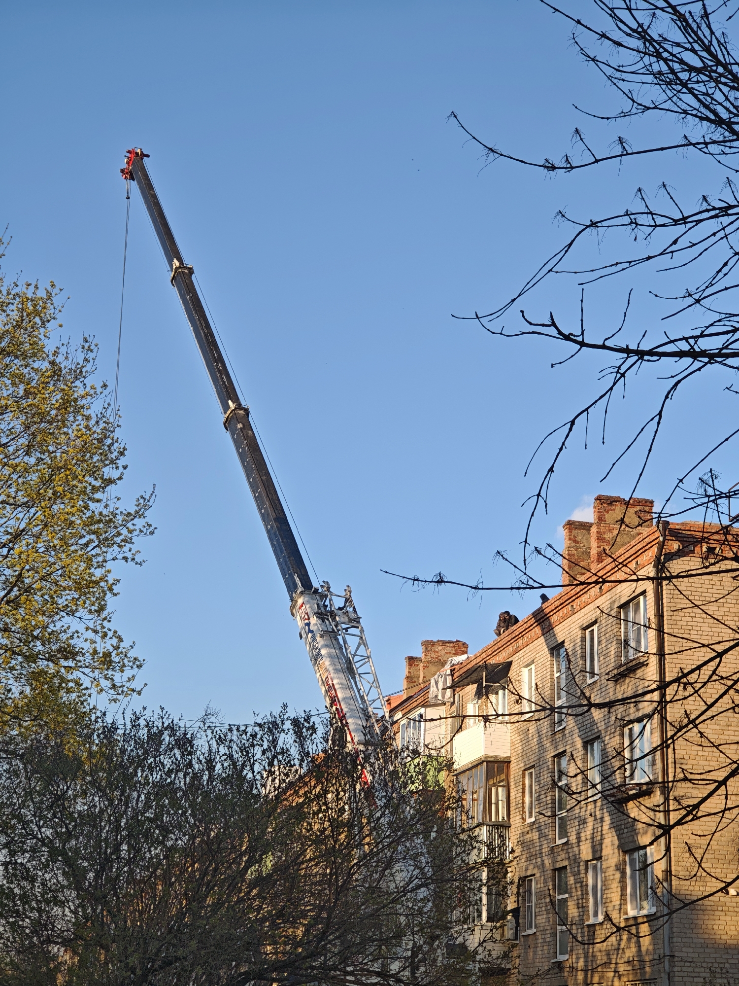 The image depicts an extended crane working on a brick apartment building. The location appears to be Noginsk, Russia, based on visual cues such as architectural style and crane lettering. The sky is a clear blue, with some trees in the foreground partially obscuring the building and crane. The apartment building is several stories high with balconies and brick chimneys. Two individuals are visible on the roof near the crane's arm.