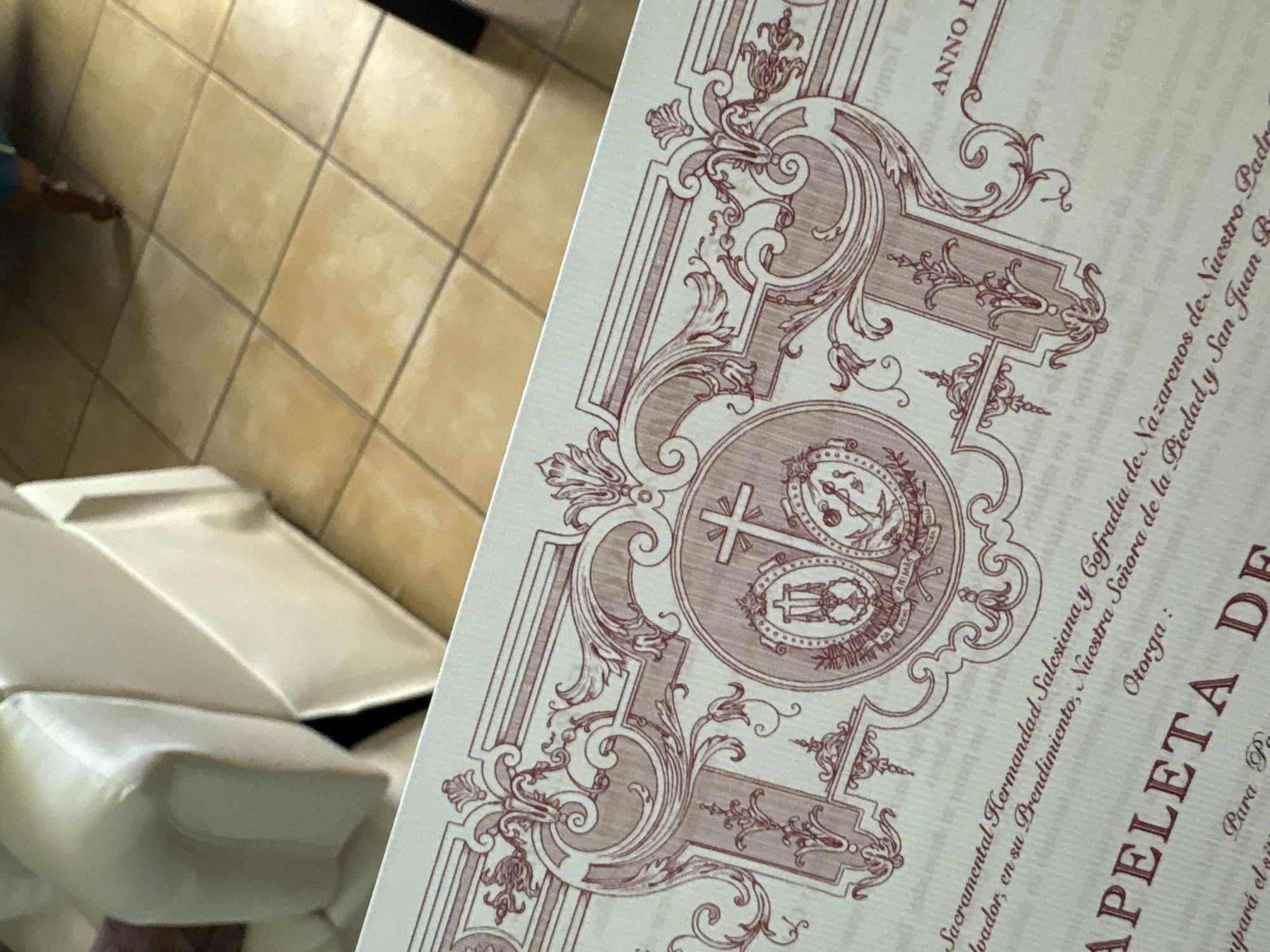 Here is a detailed description of the image:
**Scene Overview:**
The image shows a close-up view of a printed document with ornate, classical-style illustrations in a red-brown color. The document appears to be partially obscuring a scene in the background. The setting suggests an indoor environment, possibly in Urbanización Encinares De Alcolea, Spain, based on the text in the document referencing Spanish religious organizations and places.
**Foreground (Document):**
* **Content:** The visible text includes phrases like "Sacramental Hermandad Salesiana y Cofradía de Nazarenos de Nuestro Padre" and "Nuestra Señora de la Piedad y San Juan R," indicating a religious or fraternal organization and locations possibly related to the city of Urbanización Encinares De Alcolea, Spain. The words "APELETA DE" are also visible, presumably part of the document's title.
* **Design:** The document features ornate, red-brown graphics including a cross and other religious symbols. The print style is reminiscent of antique religious documents.
**Background:**
* **Object:** There are white pillows or cushions with angular shapes.
* **Environment:** A tiled wall is partially visible, the tiles are light tan or beige.