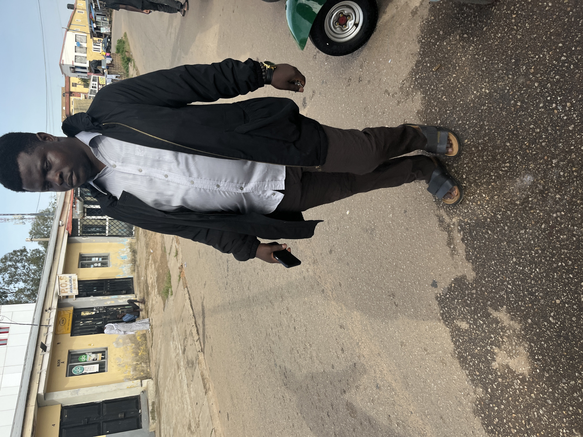 An adult male, appearing to be of Nigerian descent and dressed in a light-colored, collared shirt, dark trousers, and a black jacket, walks on a paved road in Jos, Nigeria. He is holding a dark-colored mobile phone in his right hand. The man is wearing dark sandals. To the right of the man, a portion of a green motorized tricycle is visible. In the background, a row of single-story buildings lines the street, with some displaying signage, including "POS AVAILABLE HERE" and logos for telecommunication companies. A few individuals are standing and sitting near the entrance of one of the buildings, and a white cloth is draped over what appears to be a person or object inside a doorway. The road surface shows patches of wetness and debris. The overall environment suggests a commercial or residential street in Jos.