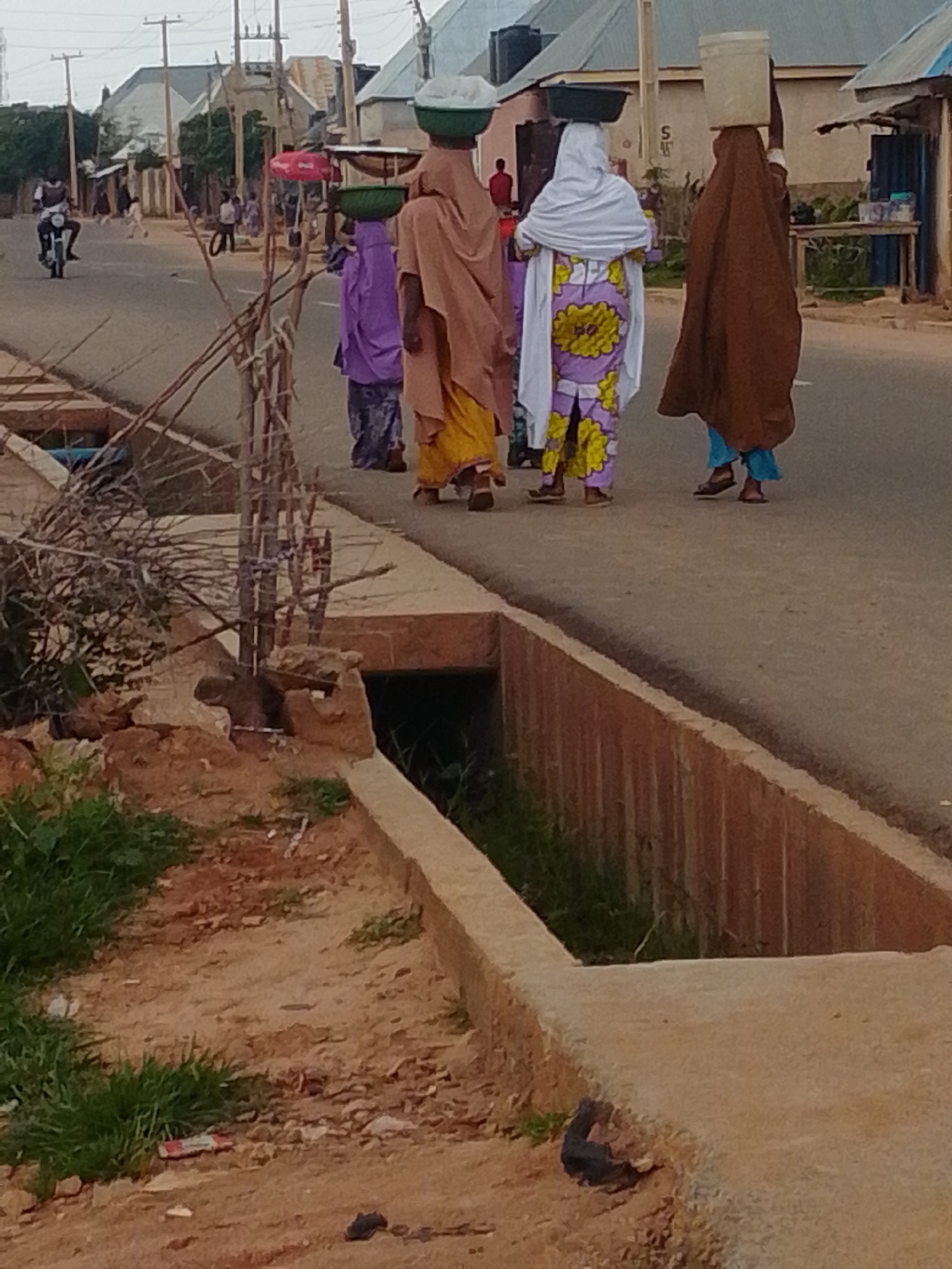 In Gombe, Nigeria, a dirt road runs through a residential area. Several women walk along the road, some carrying items on their heads. The first woman wears a light brown headscarf and a long, flowing yellow dress. Next to her, another woman wears a purple head covering and a dark skirt. The third woman from the right is dressed in a white headscarf and a purple dress with a yellow floral pattern. The last woman on the right wears a brown head covering and a blue skirt, carrying a large white bucket on her head. A motorcycle is visible on the left side of the road, with a person riding it. In the foreground, there is a drainage ditch with concrete edging and a rough, muddy embankment. Vegetation, including grass and branches, grows on the embankment. Buildings with corrugated metal roofs are visible in the background, suggesting an urban or semi-urban environment. The lighting suggests it is daytime.