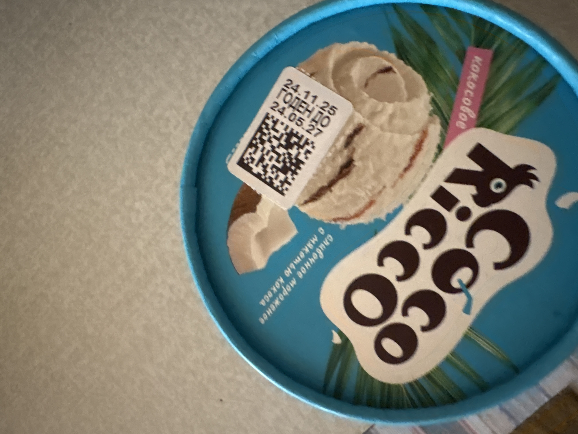 The image displays a close-up, angled view of the blue lid of an ice cream container, resting on a light, textured surface. The lid is predominantly a vibrant blue, featuring illustrations of creamy white ice cream, pieces of coconut, and green palm-like leaves.
A stylized brand name, "Ricco", is prominent on the lid in large, brown, bubble-like letters within a white, irregular shape, with "coco" emphasized as part of the design. The word "кокосовое" (kokosovoe), meaning "coconut" in Russian, is printed vertically in pink lettering on the right side of the lid, next to the green foliage illustration. Below the depicted ice cream, additional Russian text reads "сливочное мороженое с мякотью кокоса", which translates to "creamy ice cream with coconut pulp".
A white sticker is affixed to the top left portion of the lid. This sticker contains a black QR code at the bottom. Above the QR code, dates and text are printed: "24.11.25" followed by "ГОДЕН ДО" (Goden Do), which means "Best Before" or "Valid Until", and then "24.05.27". This indicates a production date of November 24, 2025, and an expiration date of May 24, 2027.
The lighting suggests an indoor setting, likely daylight or artificial light, with no specific time of day discernible. No people or activities are present in the image, which focuses solely on the product packaging.