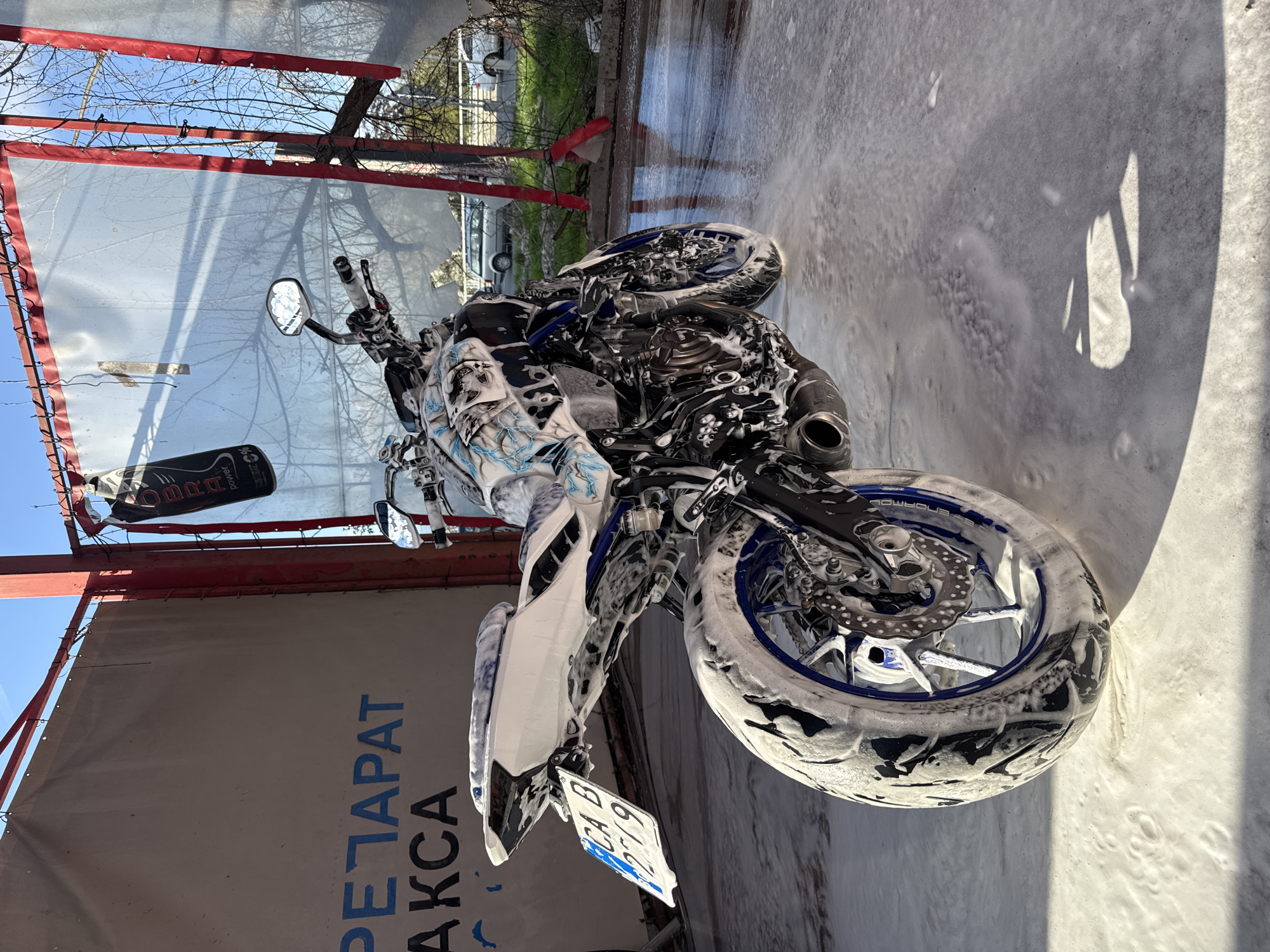 A motorcycle covered in white soap foam is being washed at a car wash. The motorcycle has a blue and white color scheme with skull graphics. Its license plate reads "CA B 2279". The car wash has a red metal frame with a translucent white covering. A sign with the partially visible text "РЕТАРАТ АКСА" hangs on the left side of the frame. The sky is clear and blue. Cars and greenery are visible in the background. The location is likely in Sofia, Bulgaria, based on the license plate. The photo is taken at an angle.