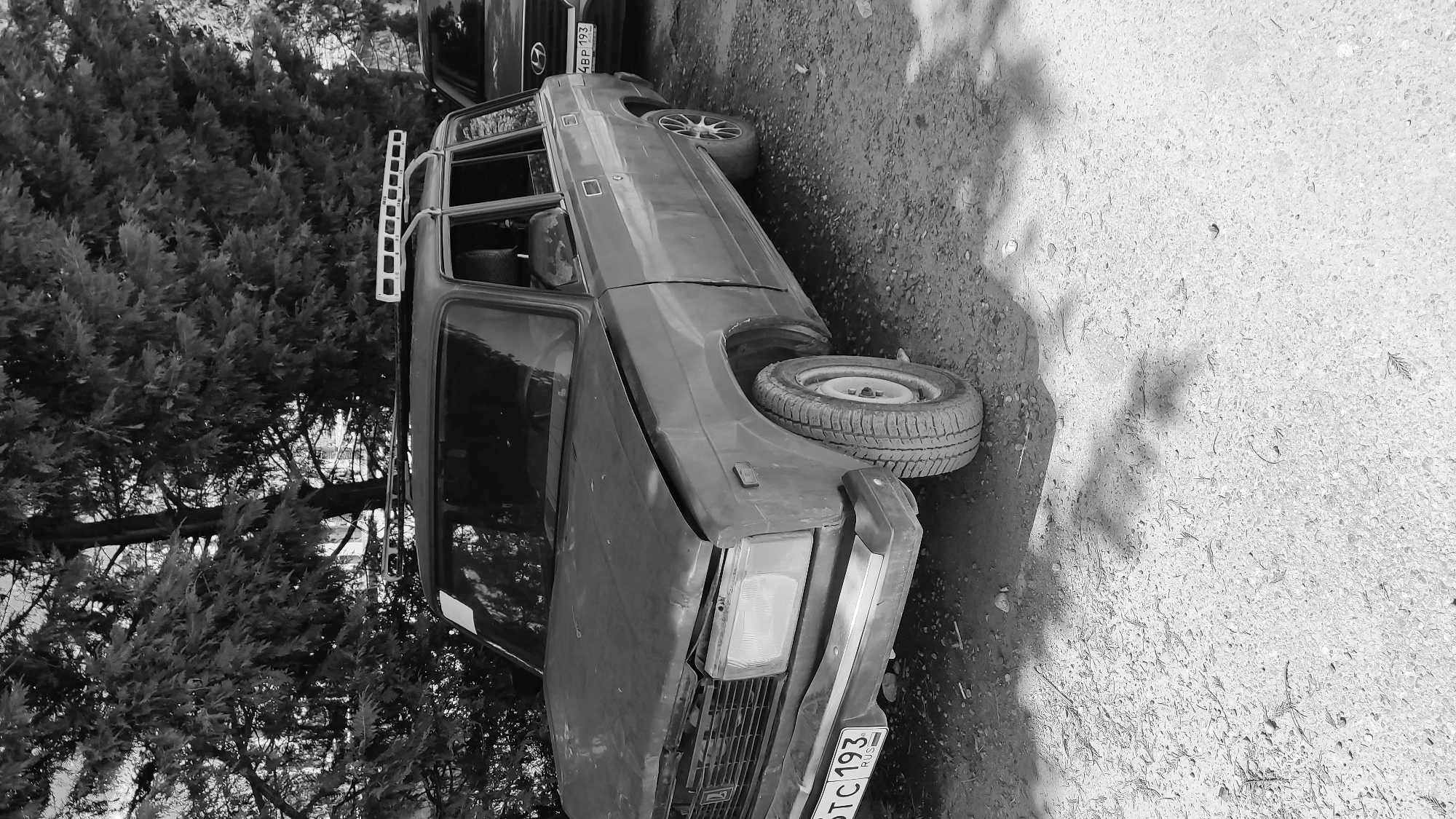 This black and white photograph captures an outdoor scene featuring two parked cars on an unpaved dirt and gravel surface, set against a dense backdrop of coniferous trees.
In the foreground, a dark-colored, older model car, likely a Lada or similar Russian make, is prominently displayed. A significant detail is the missing front passenger-side wheel, with a spare tire resting on the ground beside where the wheel should be, suggesting either a tire change is underway or the car is immobilized. The car is equipped with a roof rack. The front license plate is partially visible and reads "TC 193 RUS," indicating its origin from Russia.
Behind this car, a second dark-colored vehicle is partially visible, its own license plate showing "4BP 193," also a Russian plate. The background consists of a thick cluster of dark, possibly cypress or juniper, trees or shrubs.
The ground is uneven and appears to be a mix of dirt and small stones. Distinct shadows are cast across the ground, indicating it is daytime with clear skies. There are no people visible, and the scene is static, depicting parked vehicles. The Russian license plates, combined with the provided context, suggest the location is Krasnodar, Russia.